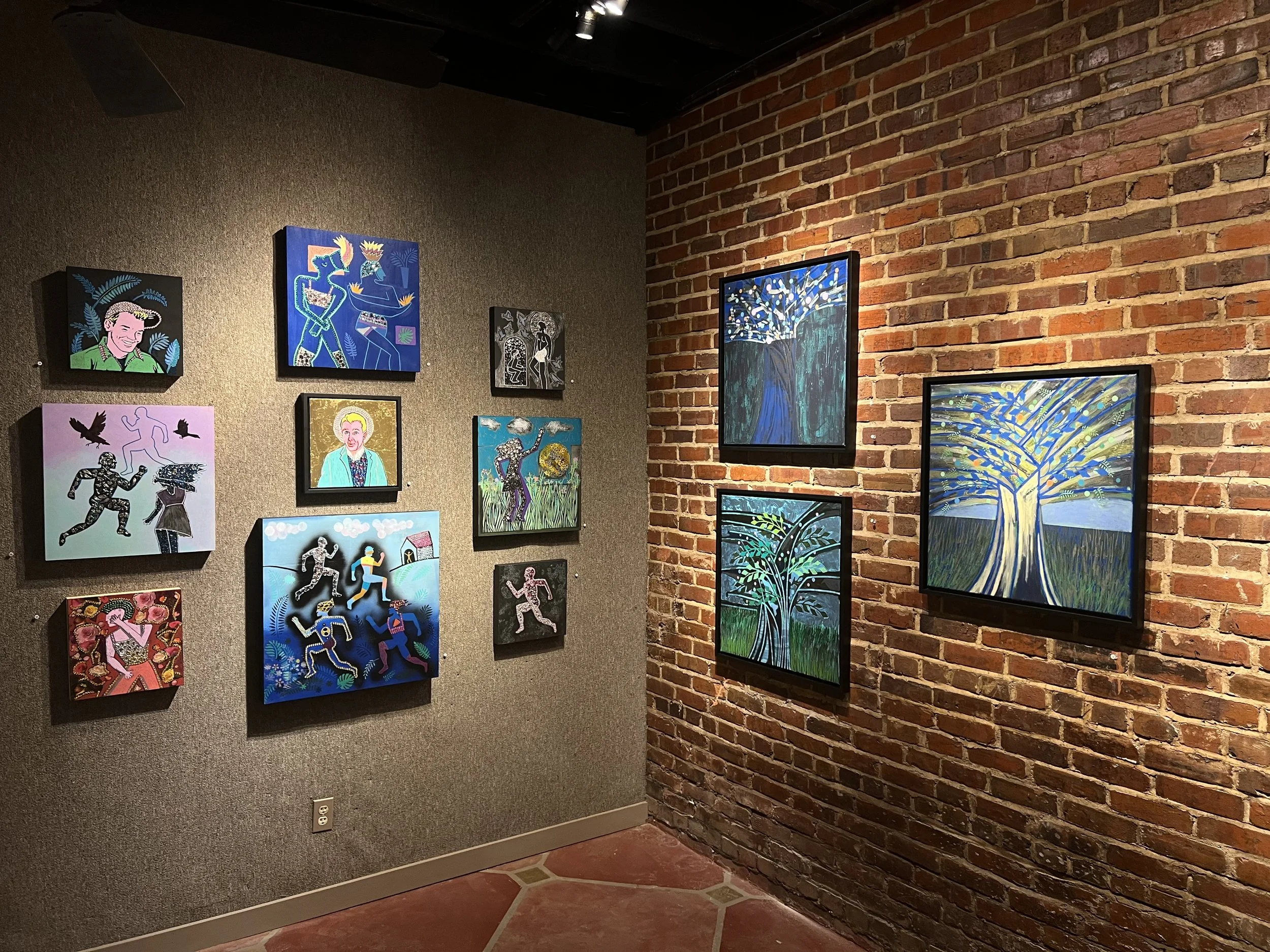 Art gallery wall with various colorful paintings, including portraits and abstract depictions of people and nature, on a wall with brown and red brick and textured gray surfaces.