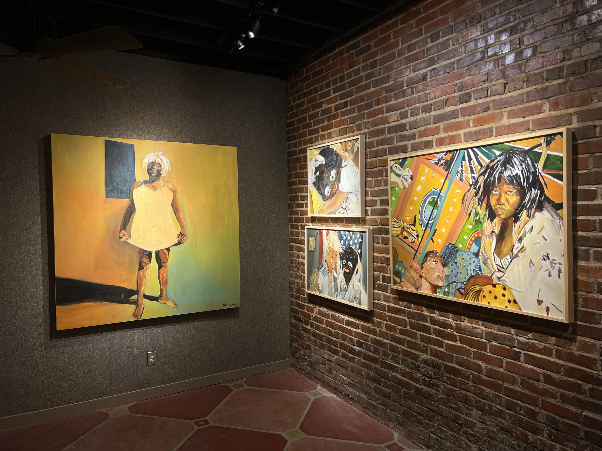 Paintings displayed on brick and grey walls in an art gallery. One of the paintings depicts a person with short white hair in a yellow dress, standing against a yellow and orange background. Other paintings show portraits and abstract scenes.
