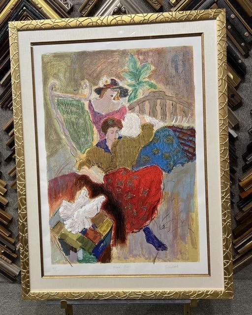 A colorful abstract painting of two women sitting outdoors, one wearing a red skirt and a large sunhat, and the other wearing a blue skirt and a purple top, with a background of trees and a bridge.