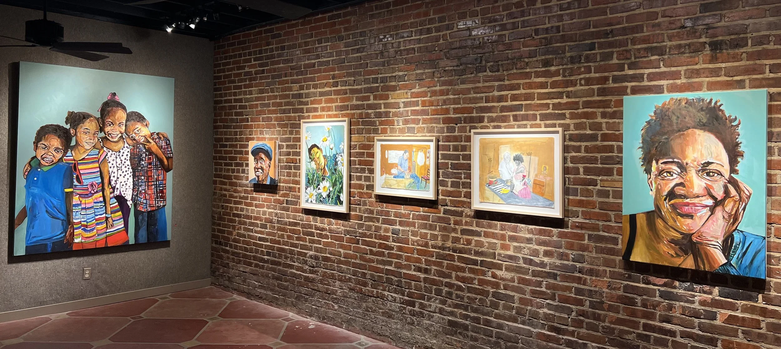 An art gallery featuring a brick wall with several colorful paintings, including portraits and landscapes. There is a large painting of four children smiling on the left, and two large portraits of women on the right side of the wall.