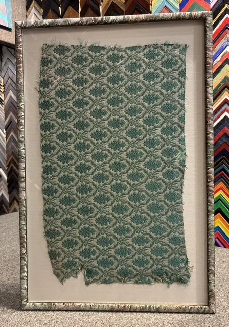 An antique fabric with a repeating geometric pattern in shades of green and beige, displayed in a frame at a fabric store.