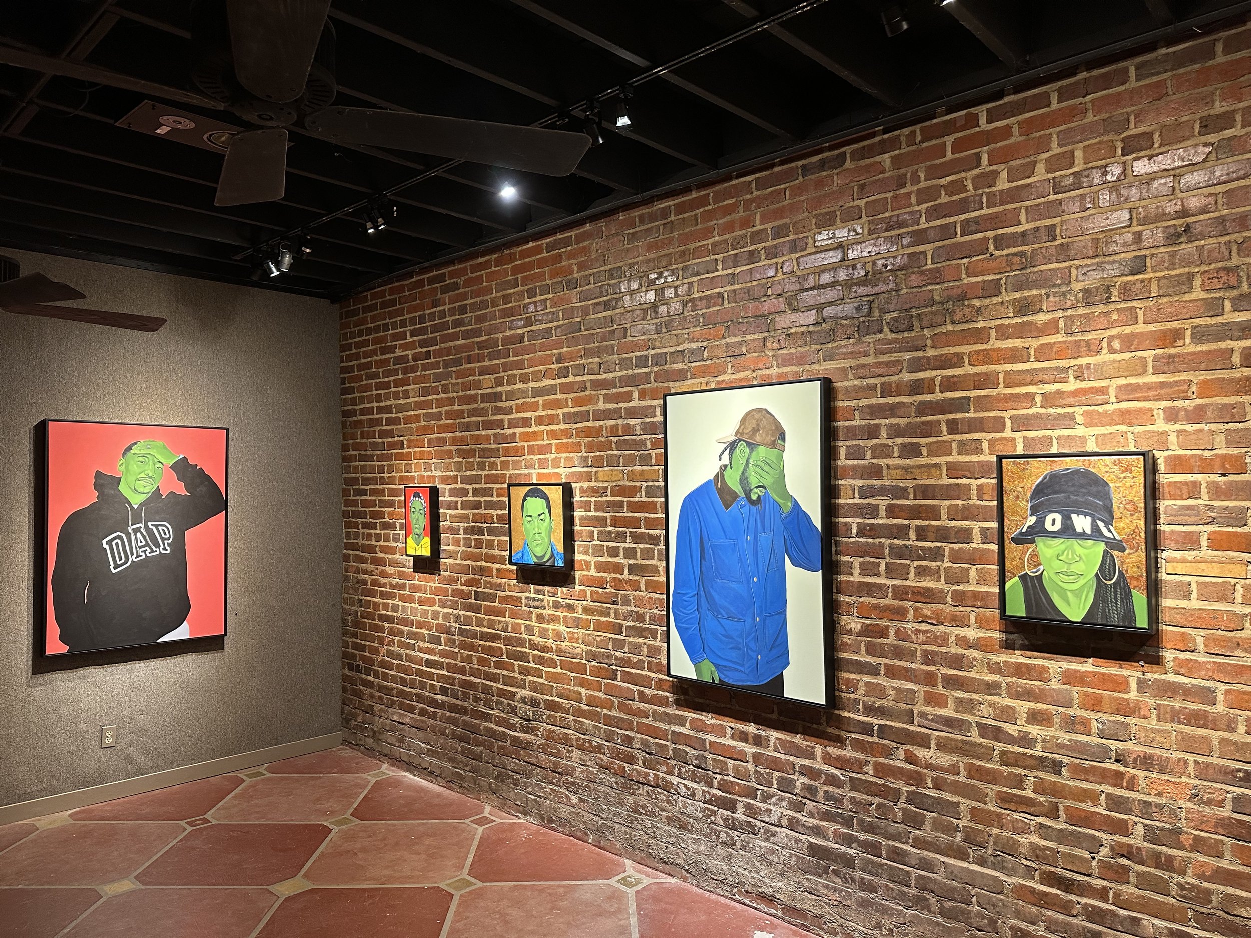 Art gallery with murals of colorful portraits of diverse individuals on a brick wall, ceiling fans, and track lighting.