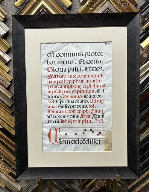 Framed illuminated manuscript page featuring Latin text in black and red ink with decorative capital and musical notation at the bottom.
