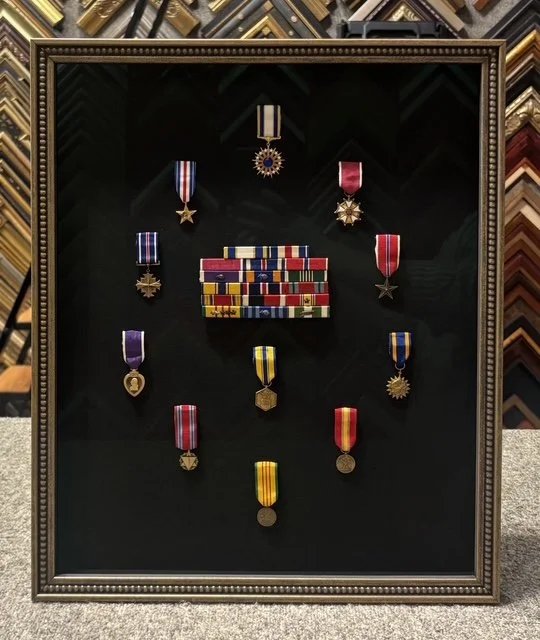 Display case with military medals and ribbons arranged on a black background inside a wooden frame, with various colorful medals and ribbons.