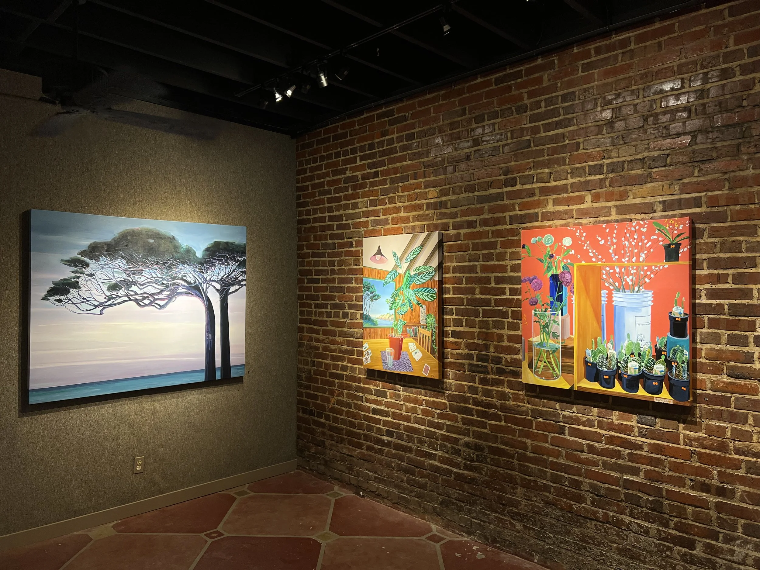 Art gallery wall displaying colorful paintings of plants and nature scenes against brick and textured walls.