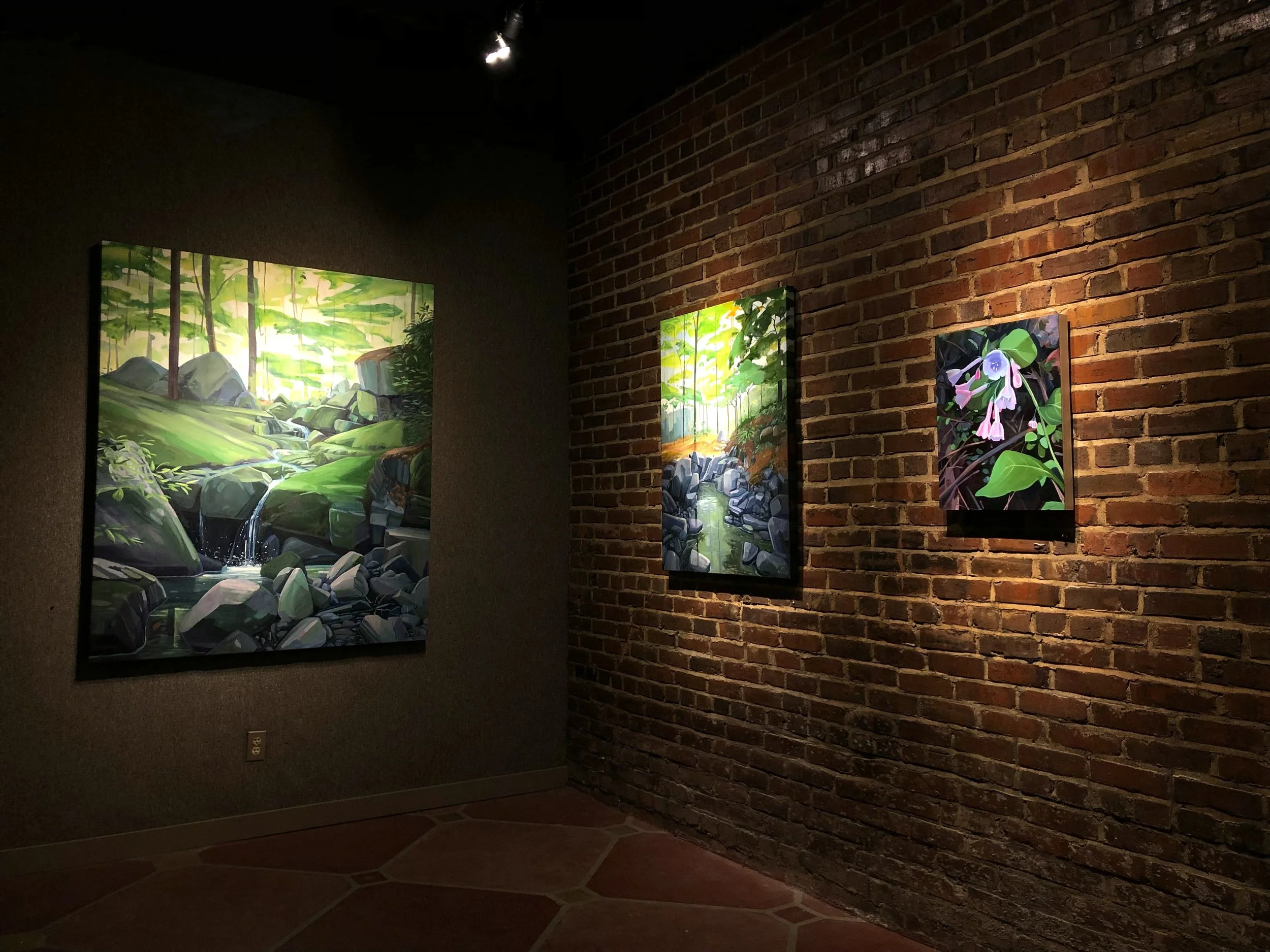 Gallery with paintings of nature scenes, including lush green forests and waterfalls, displayed on a brick wall with overhead lighting.