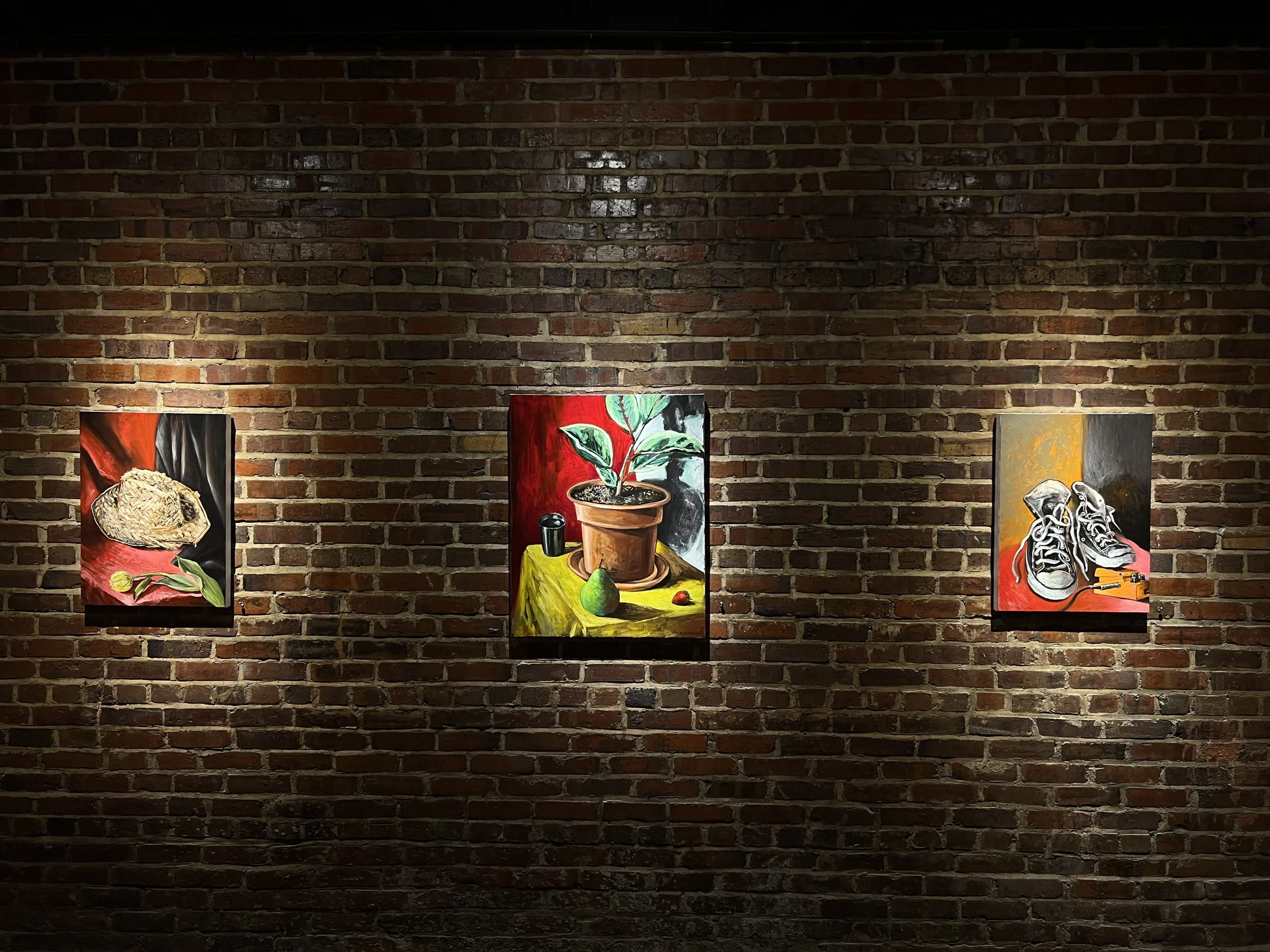 Three colorful paintings are displayed on a brick wall in a gallery. The paintings depict various objects, such as a straw hat and shoes, with abstract and realistic elements.