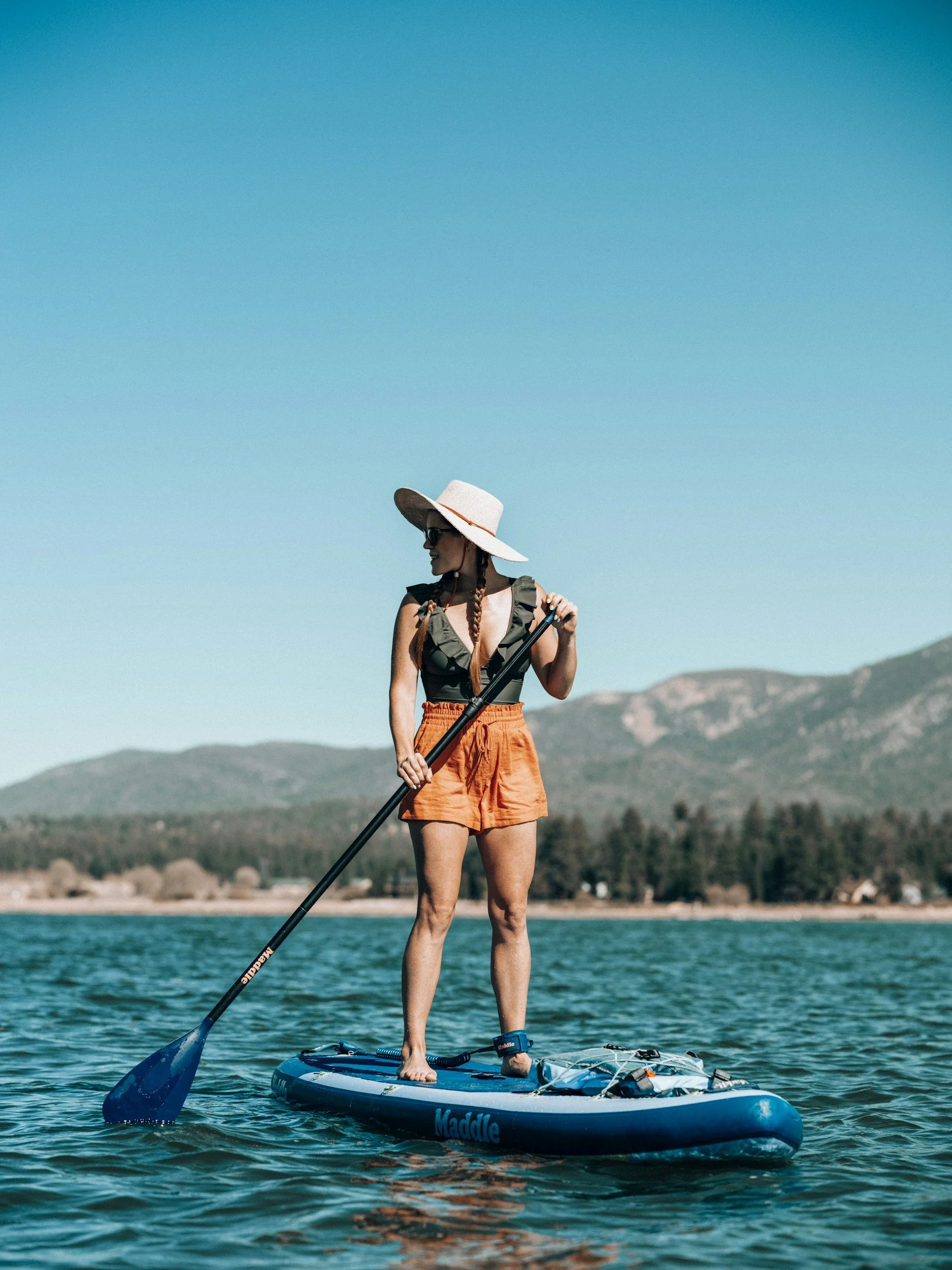 A woman in a wide-brimmed hat and sunglasses paddleboarding on a lake with mountains in the background.