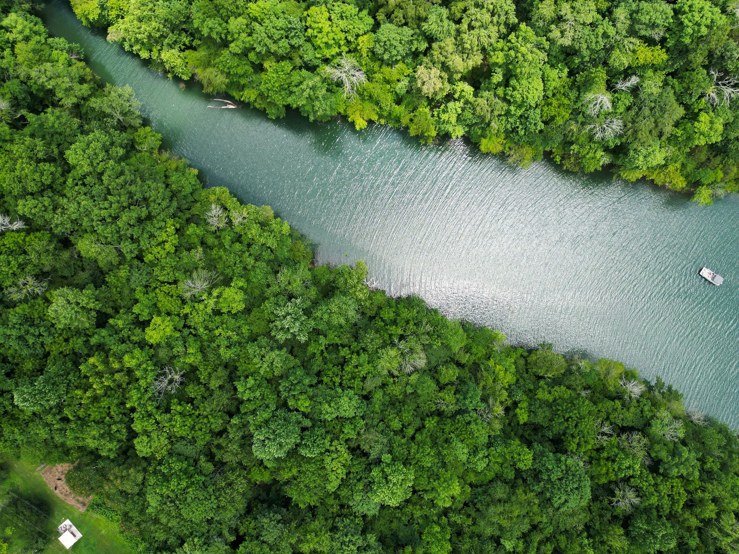 An aerial view of a river flowing through a dense green forest with a small boat near the bottom right corner.
