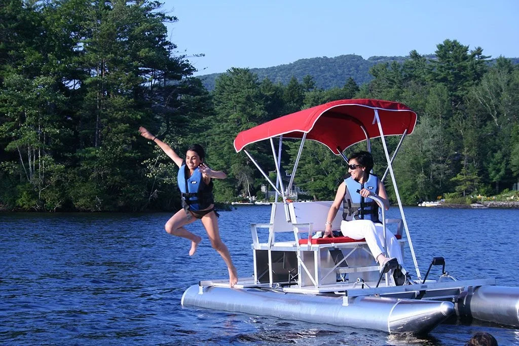 Two women on a small boat in a lake, one jumping into the water and the other sitting under a red canopy, surrounded by trees and mountains.