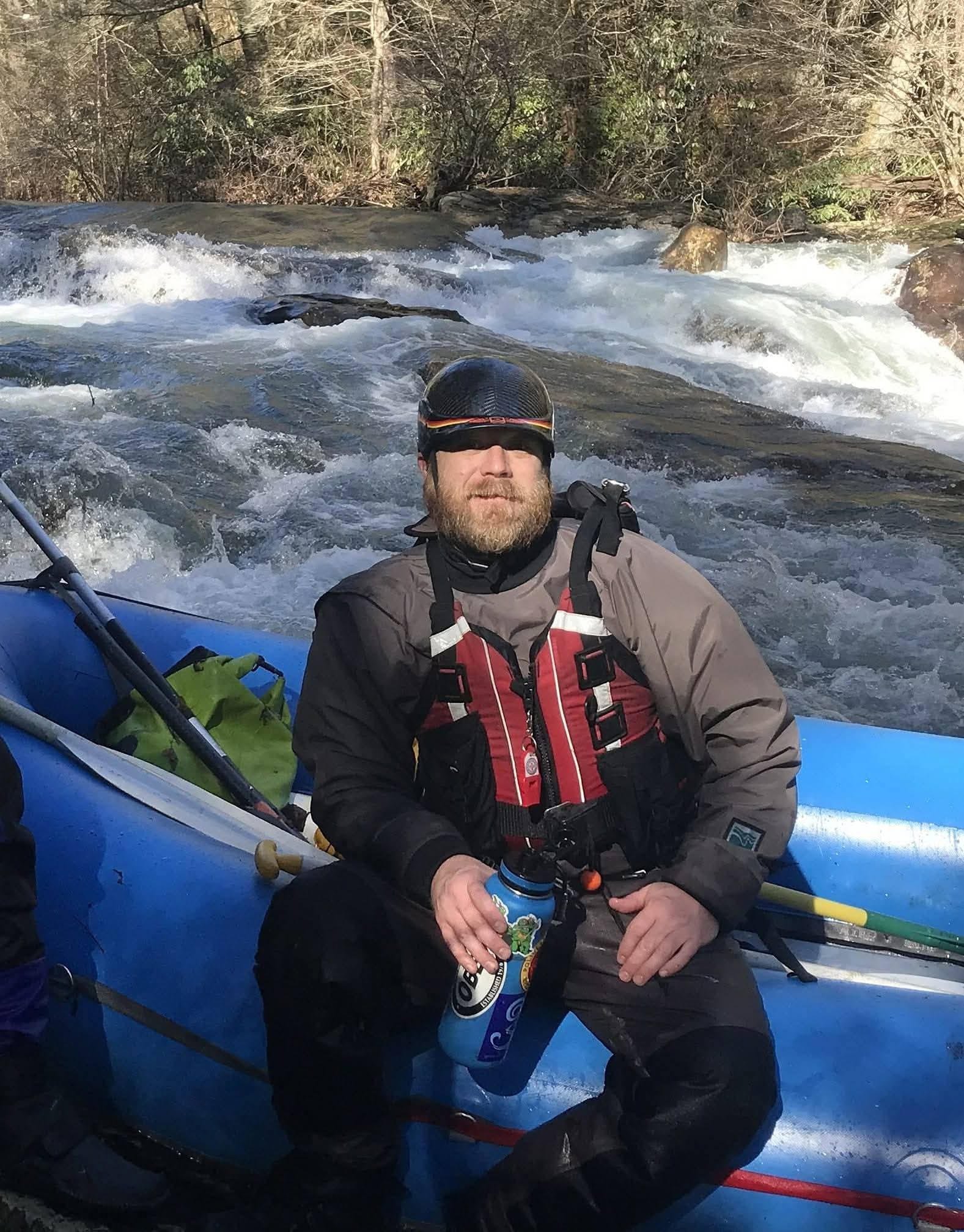 A man in rafting gear, wearing a helmet and life vest, sitting in a blue raft on a river with rapids, holding a water bottle, surrounded by forested area.