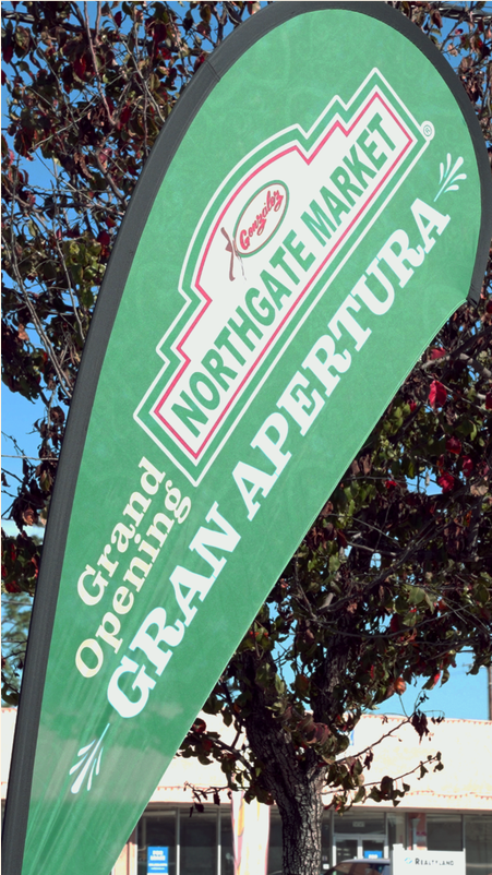 Green flag with white text reading "Grand Opening" and "Gran Aperatura" in front of a tree and storefront.