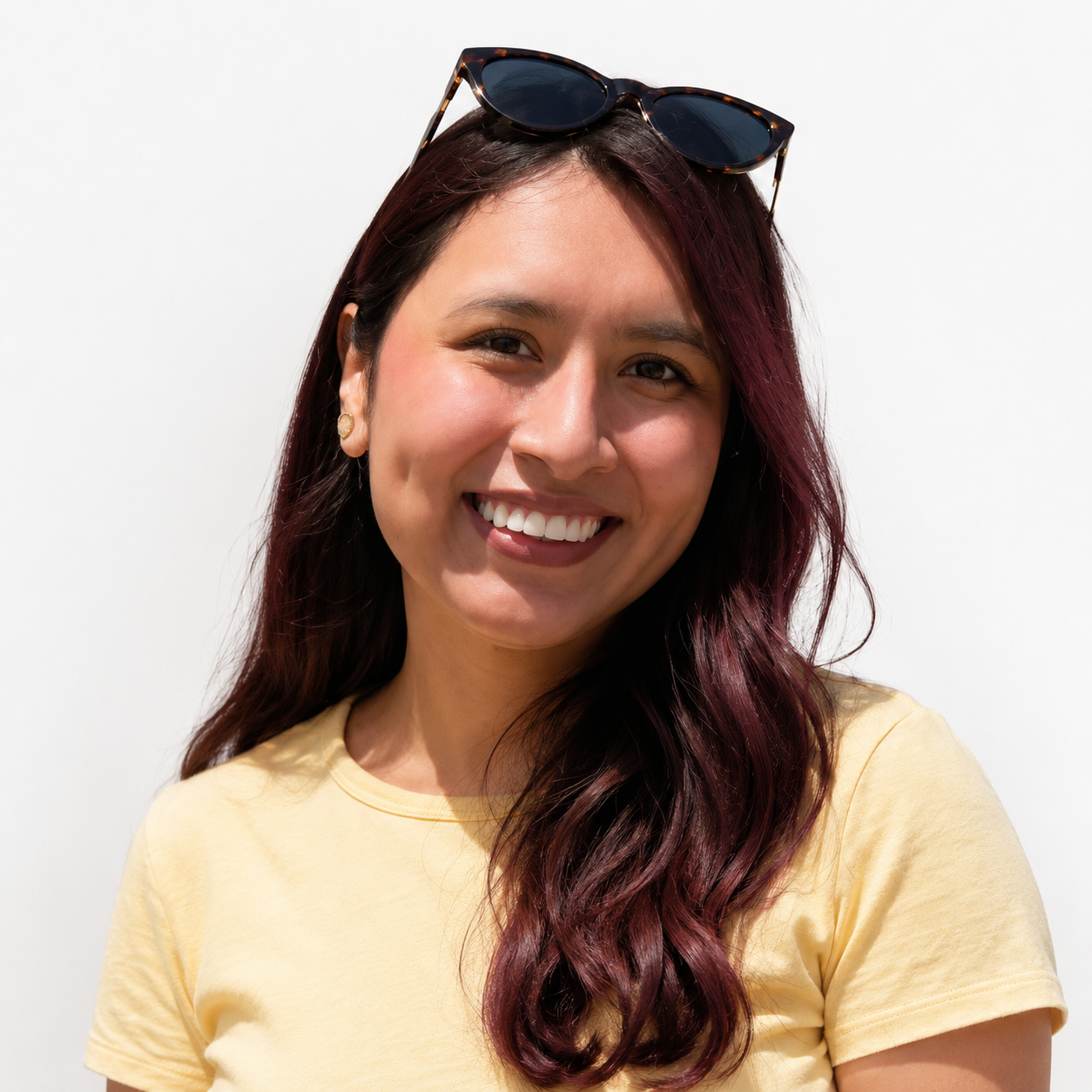 A Hispanic woman with long wavy hair, wearing a yellow t-shirt, smiling with sunglasses on her head, and earrings, against a plain white background.