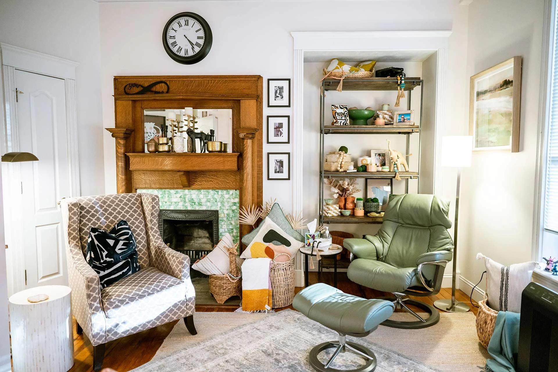 Living room with a fireplace, a wooden mantel, a wall clock, bookshelf, armchair, ottoman, and various decorative items and pillows.