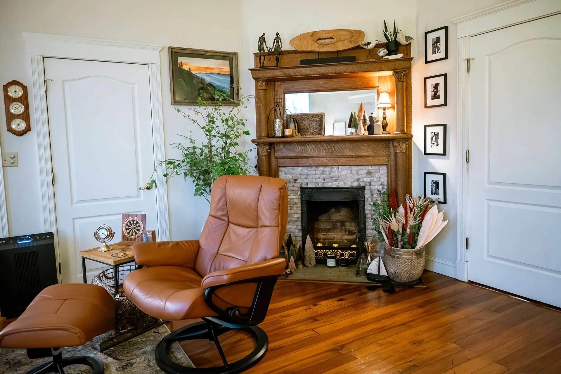 Living room with a leather armchair in front of a fireplace with a mirror, decorated with plants, artwork, and figurines.