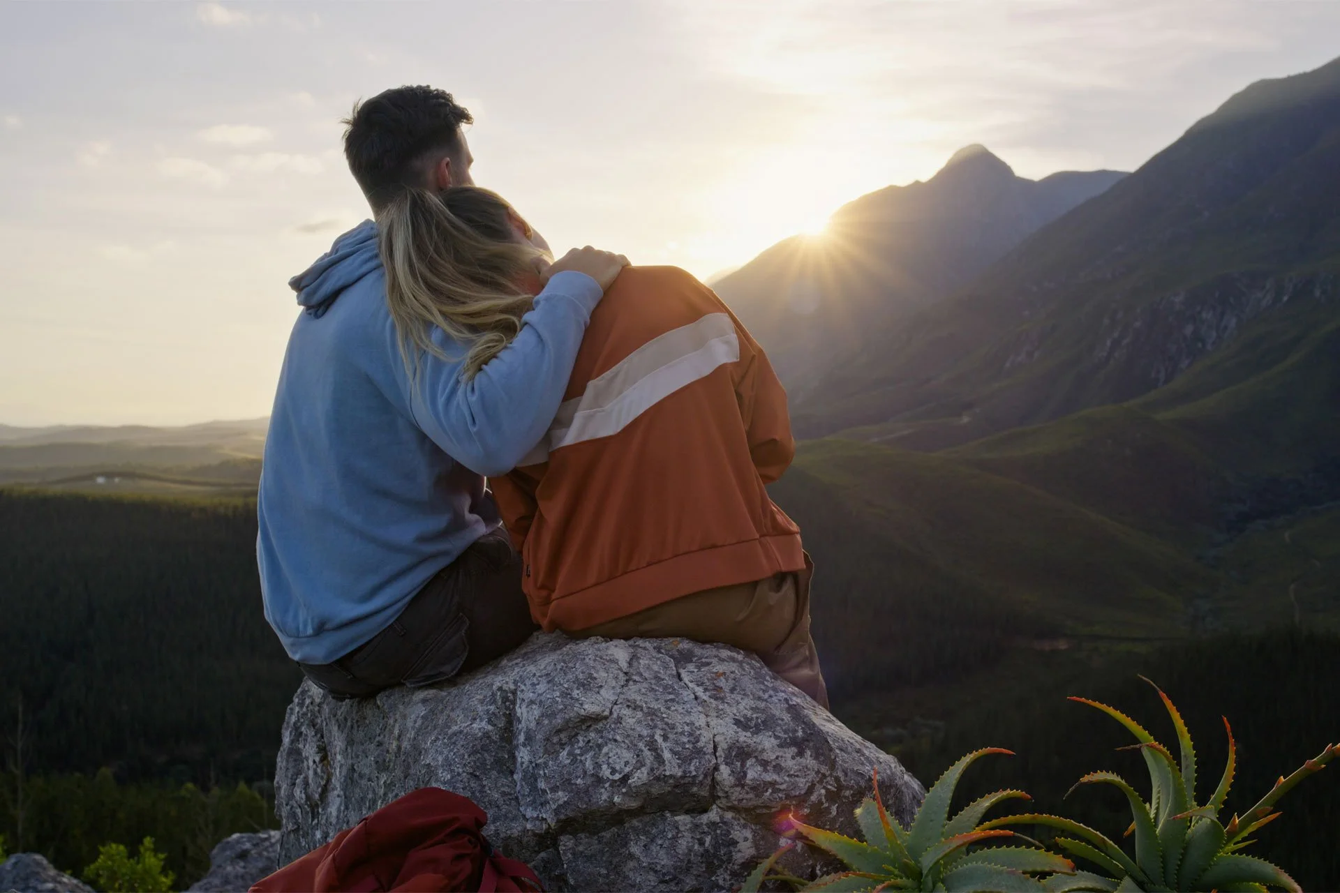 A couple sitting on a rock, embracing and watching the sunset over a mountainous landscape with green hills and a partly cloudy sky.