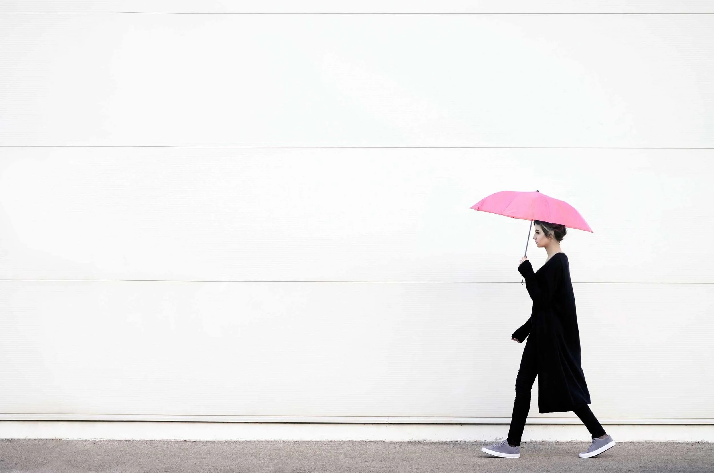 A young woman walking along a sidewalk holding a pink umbrella. She is wearing a long black coat, black leggings, and gray sneakers. The background is a white horizontal panel wall.