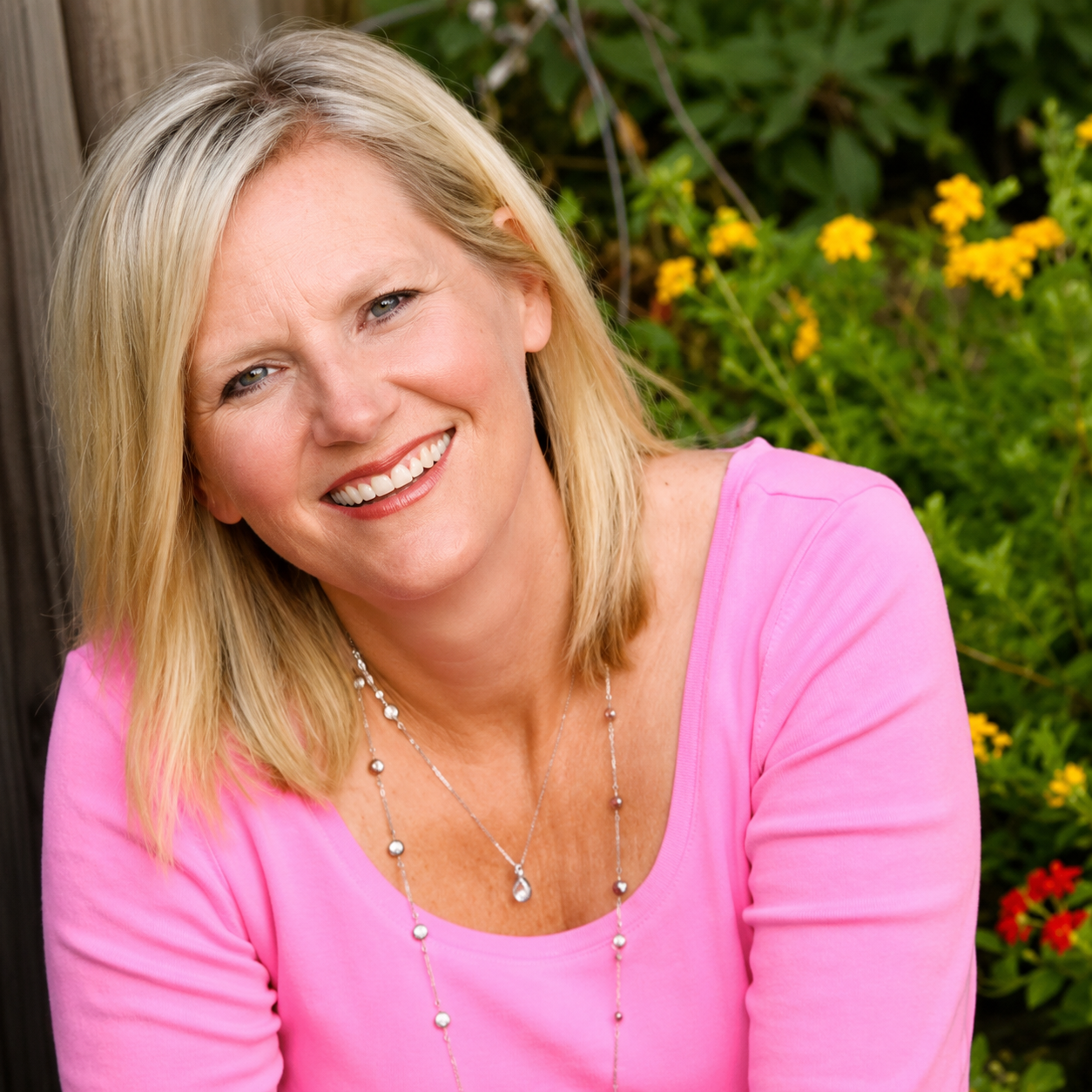 A smiling blonde woman wearing a pink top and jewelry, sitting outdoors with yellow flowers and green plants in the background.