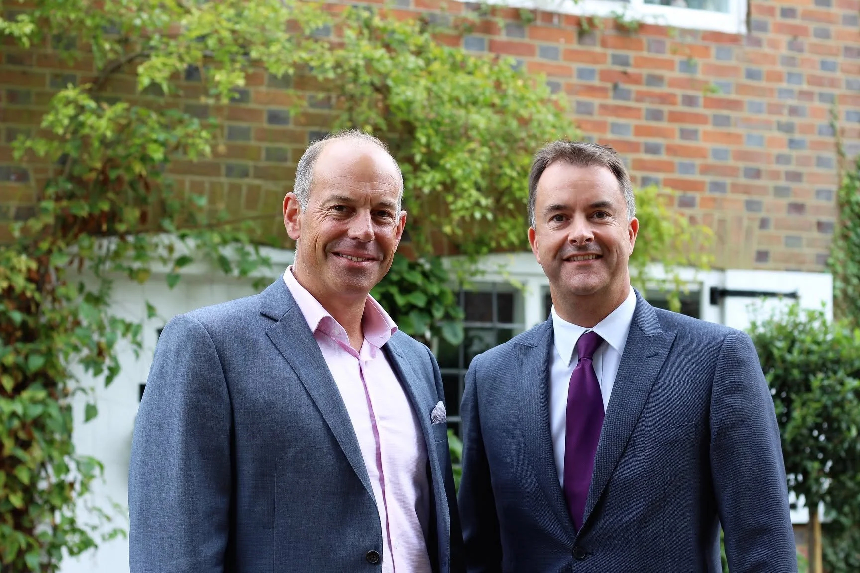 Two men dressed in suits standing outside, smiling, with a brick house and green foliage in the background.