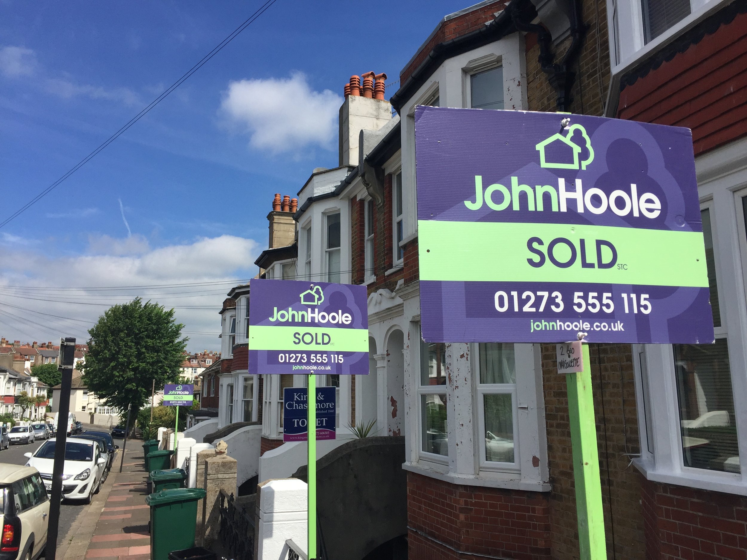 Real estate signs on a sidewalk outside houses, indicating properties are sold. The signs feature a purple and green color scheme and display the name 'John Hoole' along with contact information.