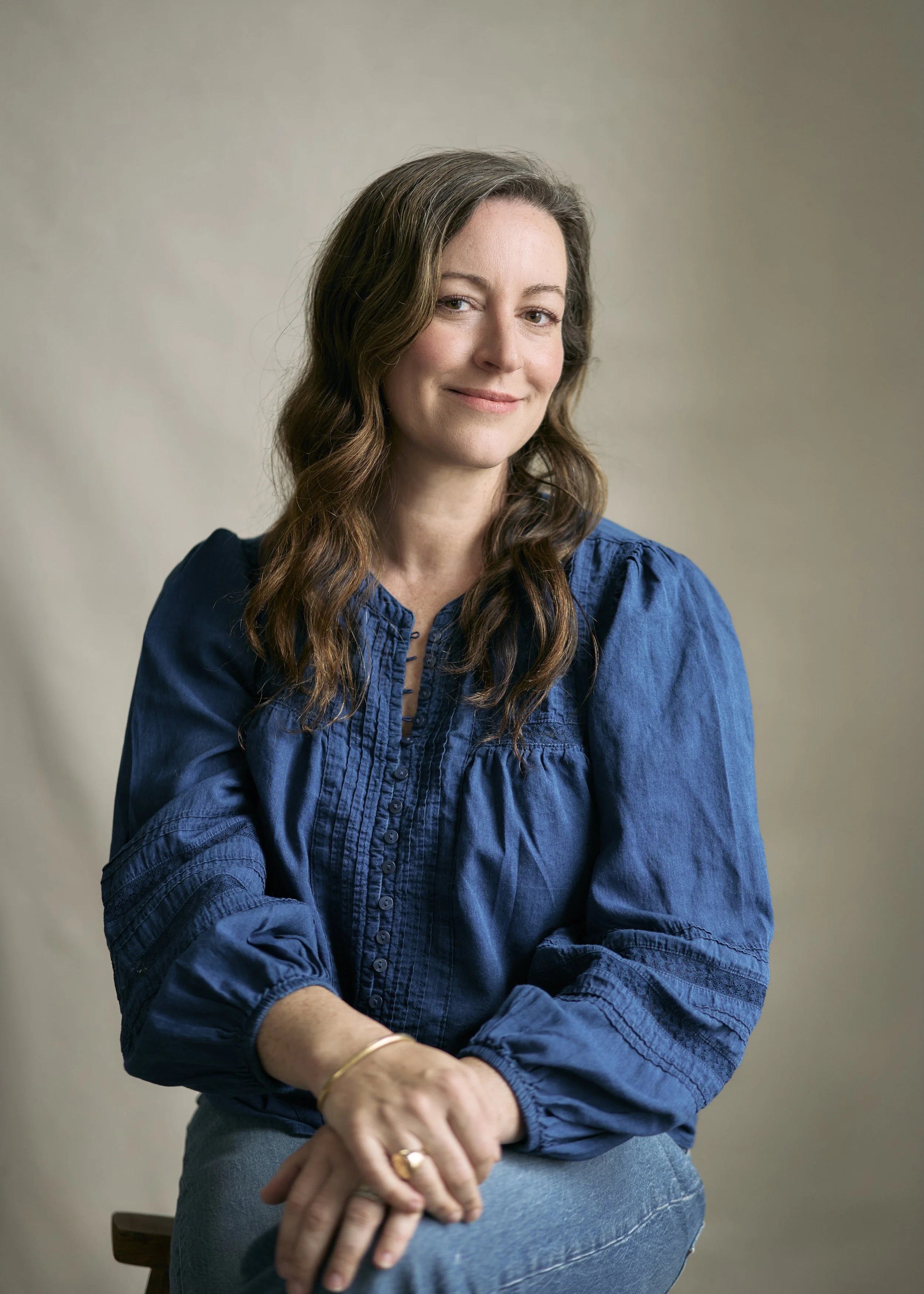 A woman with long brown hair and light skin, wearing a blue blouse with ruffled details, sitting on a wooden chair, smiling slightly, with an unfocused neutral background.