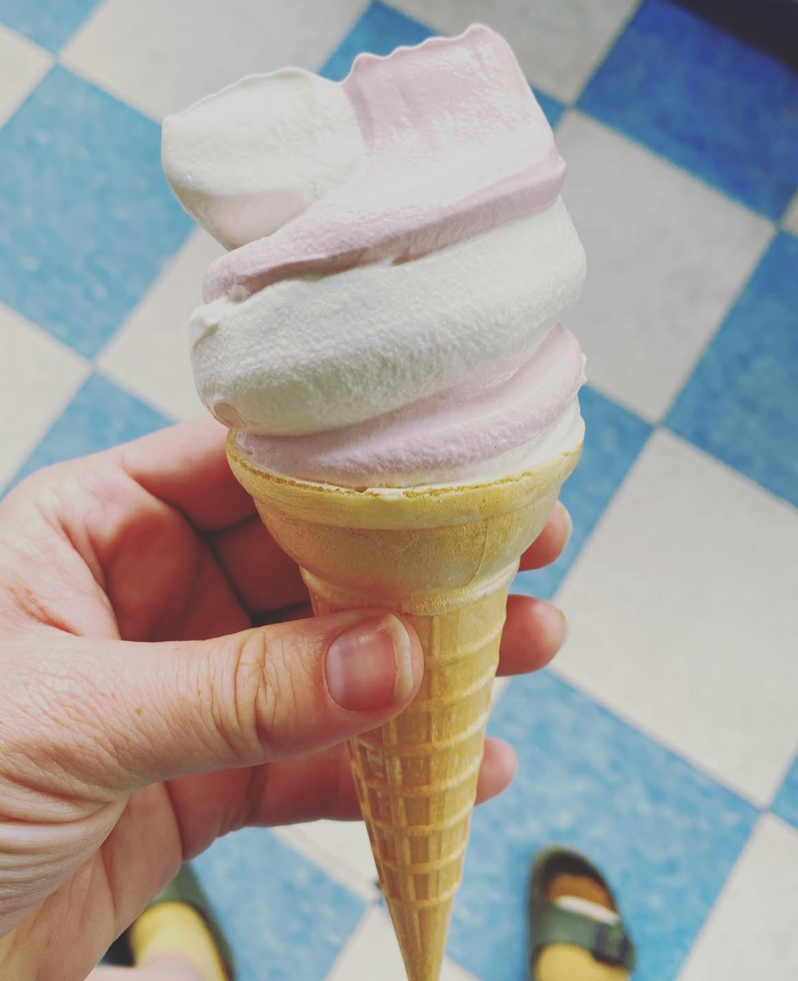 A person holding a swirl of soft serve ice cream in a waffle cone, with a background of blue and white checkered floor tiles.