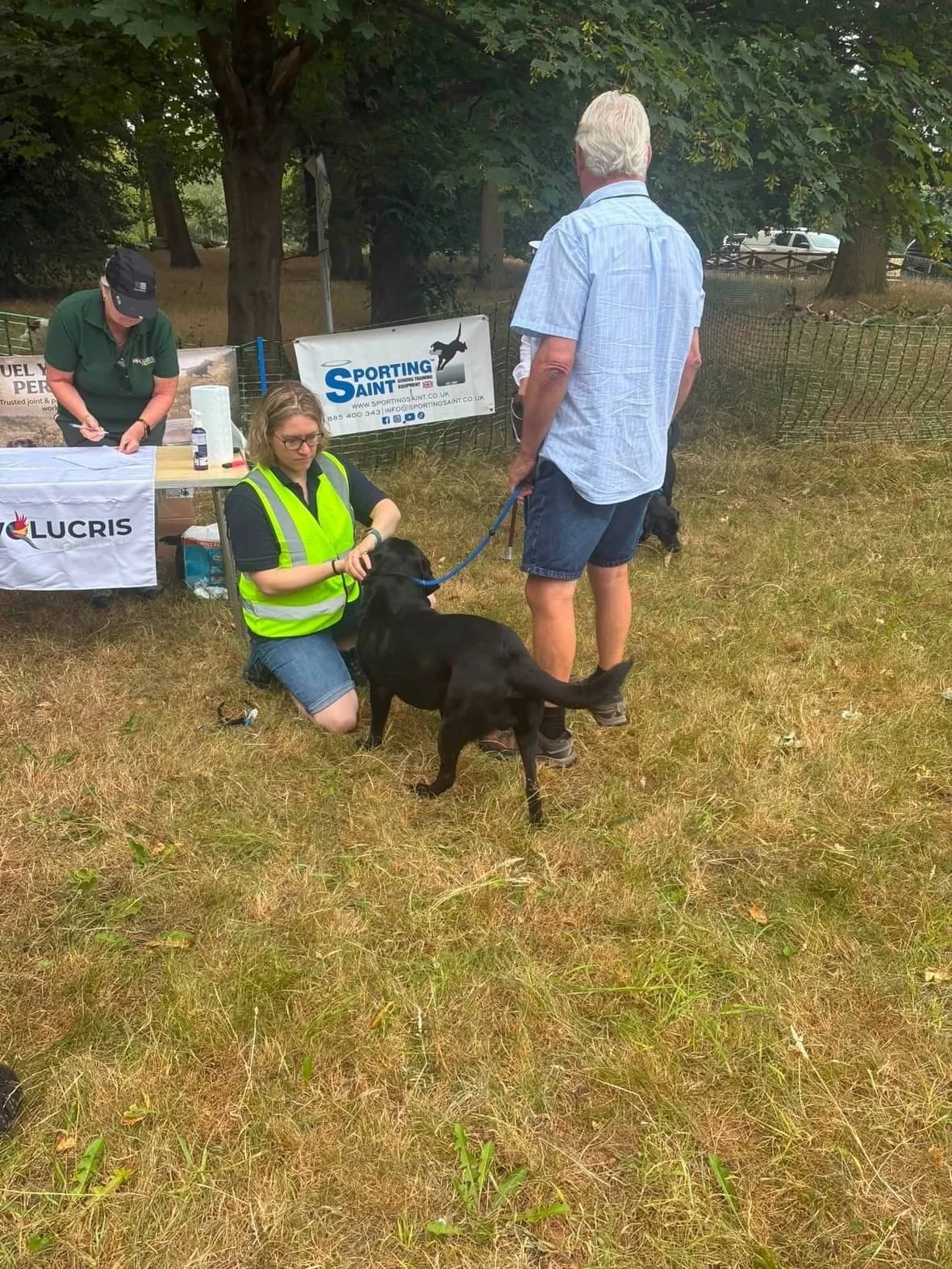 A man and a woman with a black dog at an outdoor event, with a table and banners in the background.