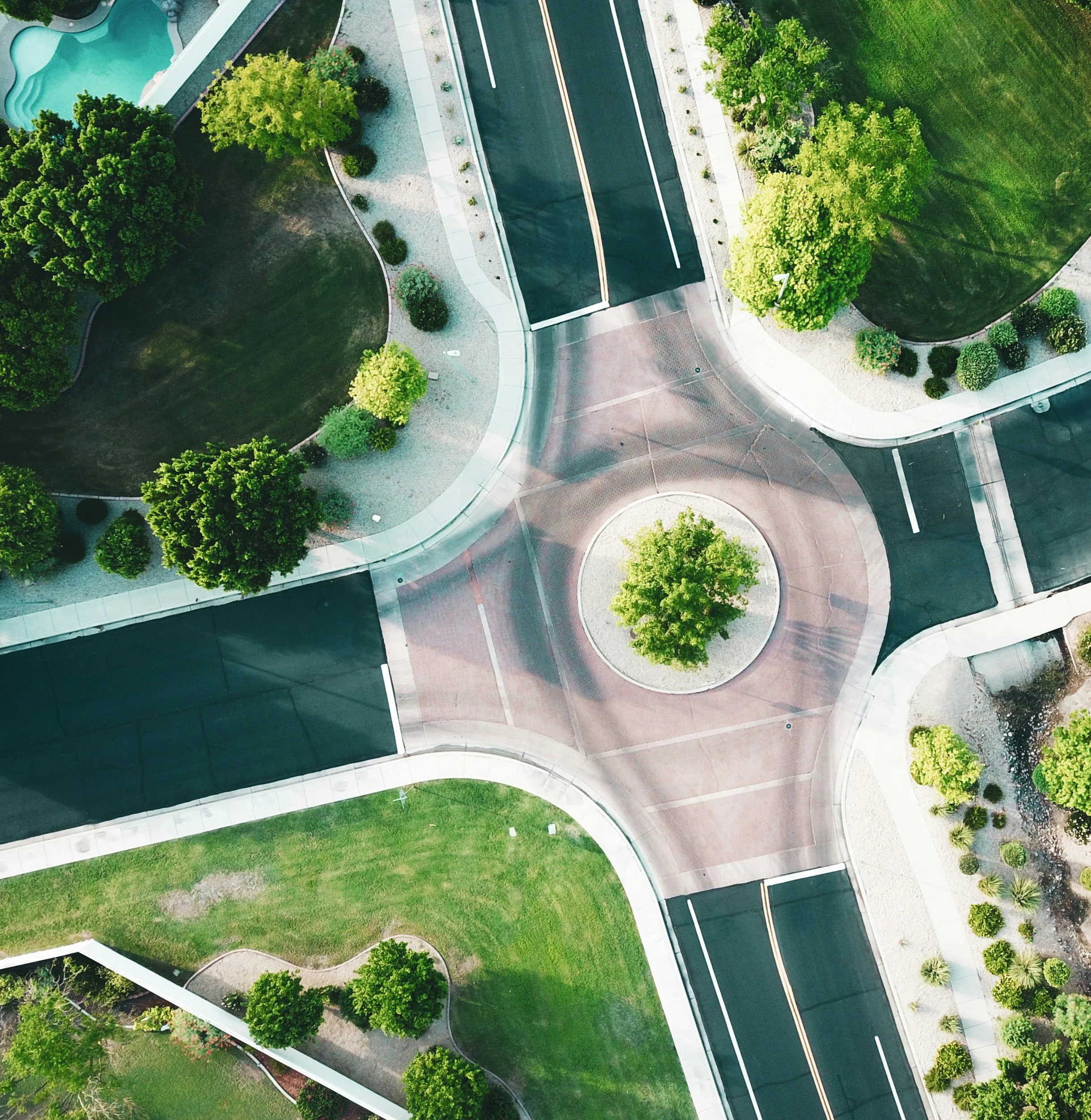 Aerial view of a neighborhood with trees, grassy areas, and a winding road with yellow dividing lines, alongside other roads and sidewalks.