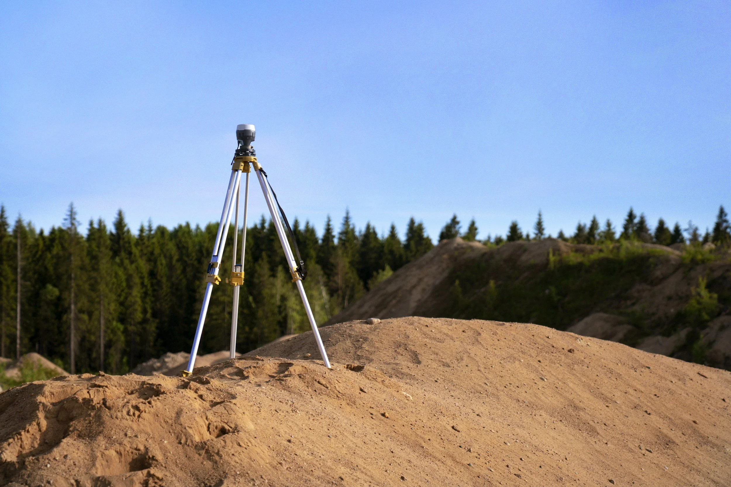 A surveyor's tripod stands on a dirt hill under a clear blue sky, with a forest in the background.