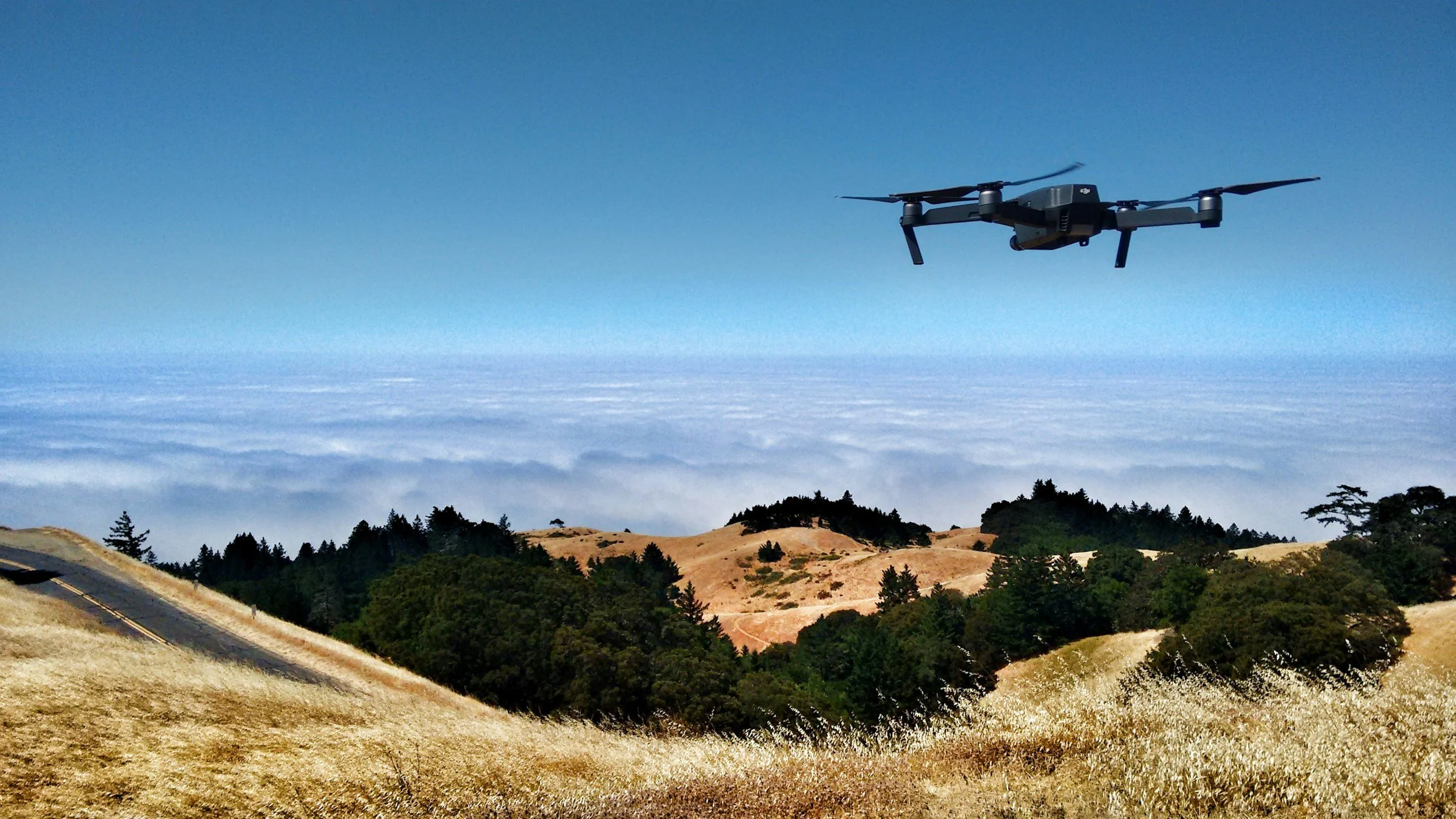 A drone flying over a landscape with rolling hills, trees, and a cloudy sky.