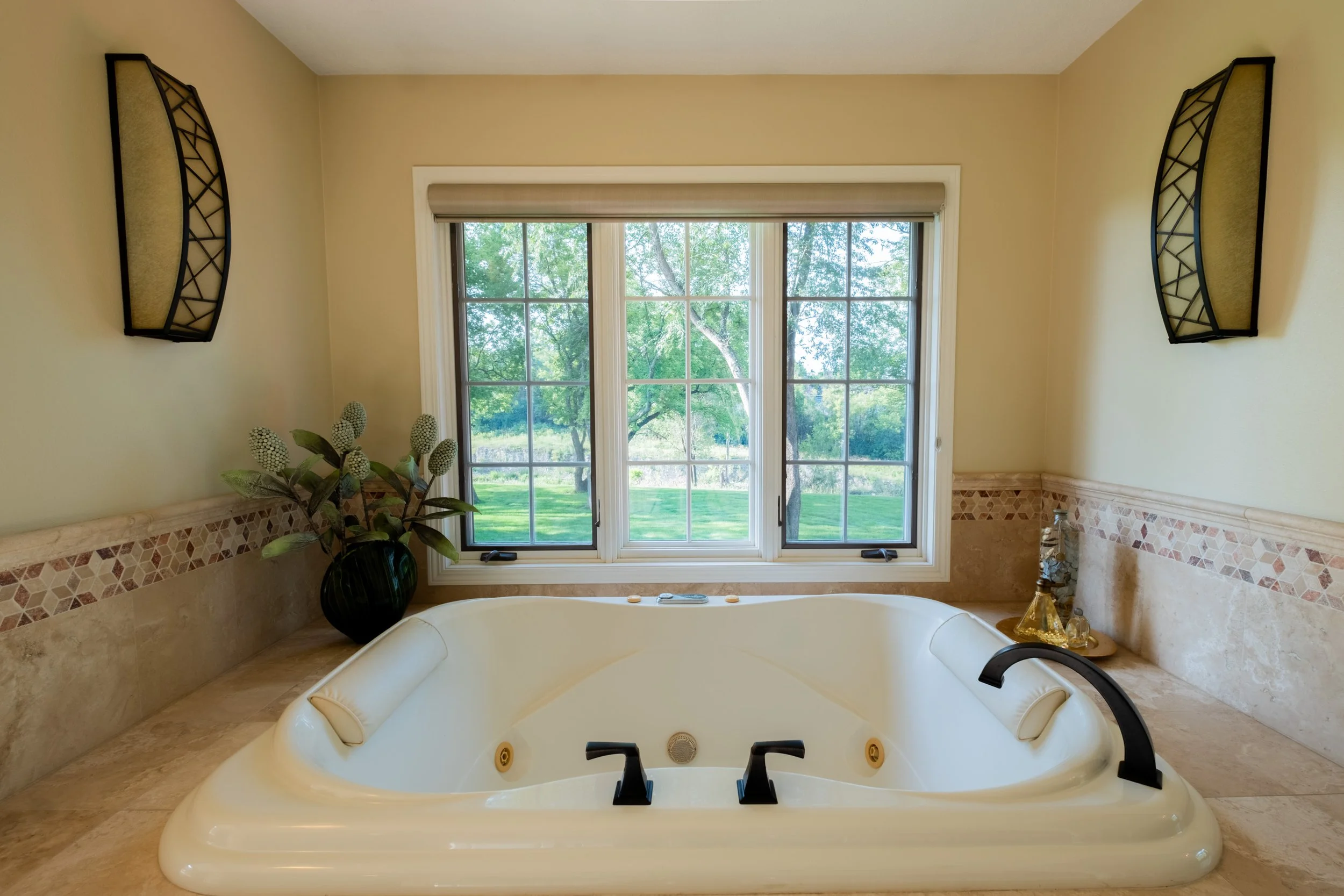 A bathroom with a large window showing green trees outside. There is a bathtub in the foreground, a potted plant on the left, and wall sconces on each side of the window. Items are placed on the ledge near the window.