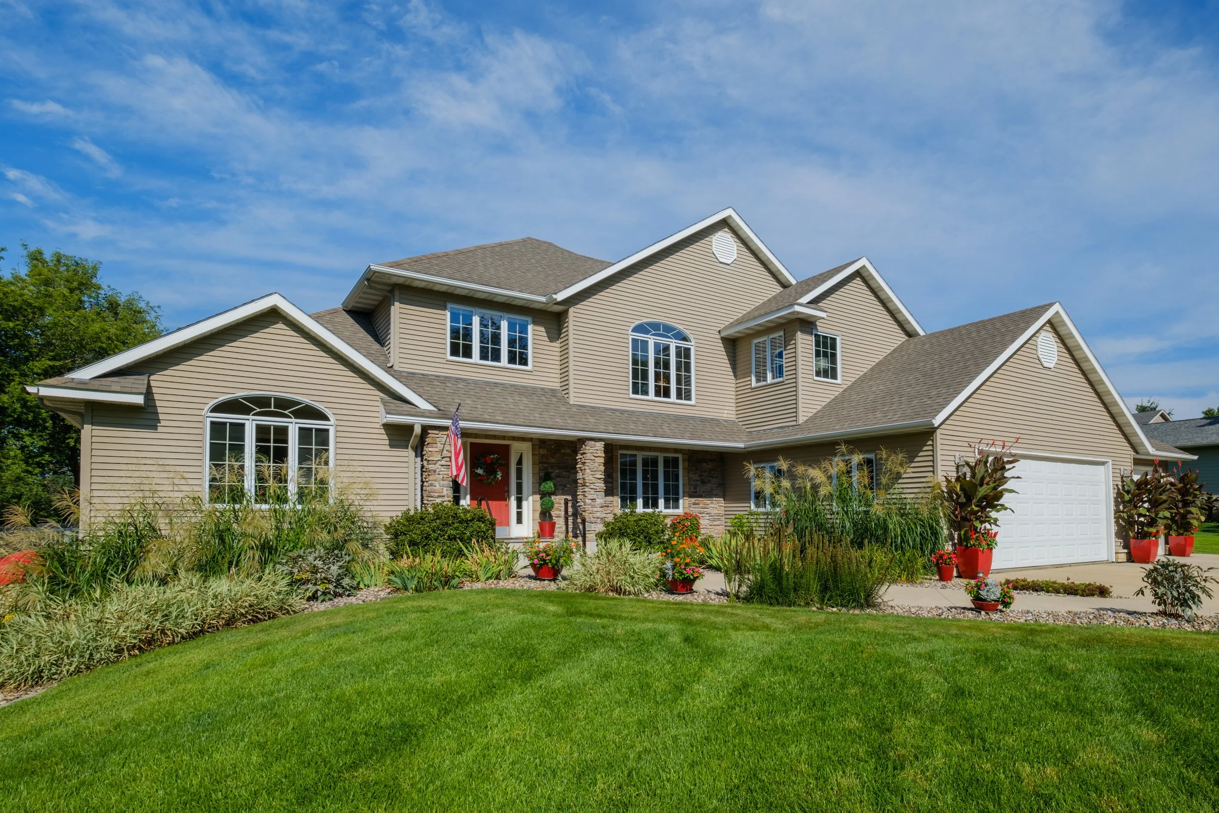A two-story suburban house with beige siding, white trim, and a gray shingled roof. The house has multiple large windows, a front porch with stone columns, and a white garage door. The front yard features a well-maintained lawn, various green plants,