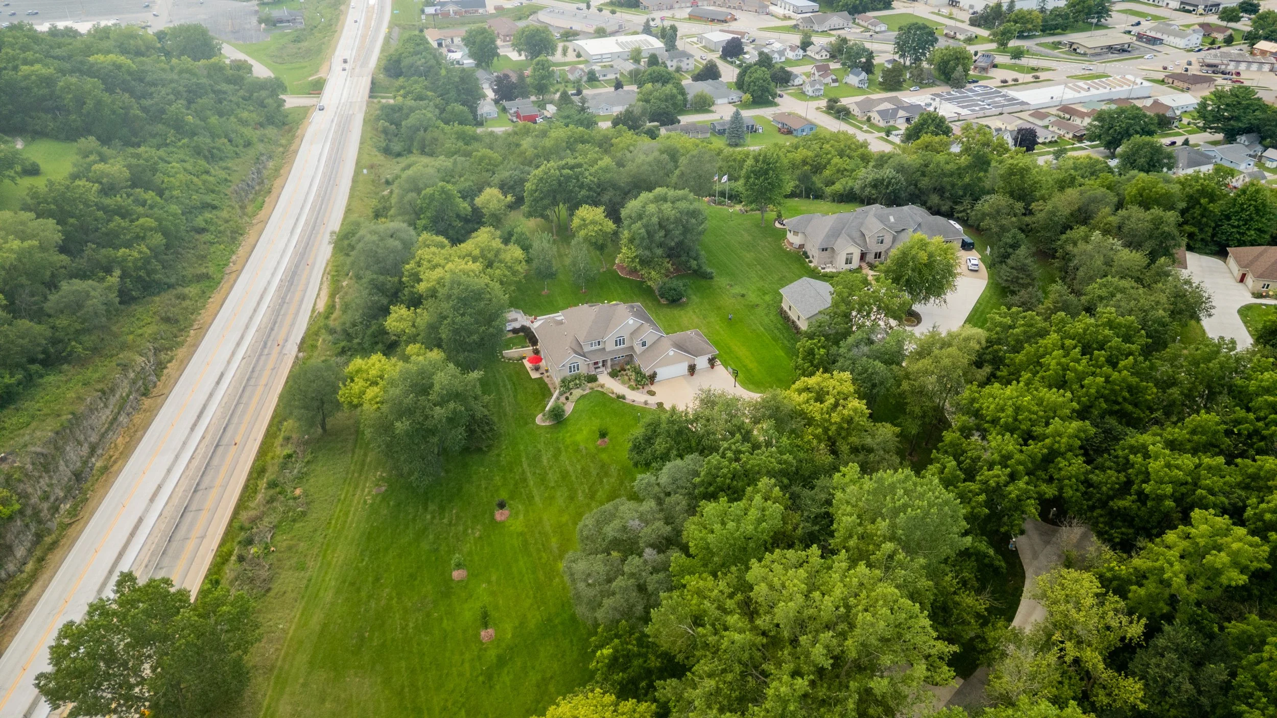 Aerial view of a suburban neighborhood with houses, lush green trees, and well-maintained lawns, with a highway running along the edge of the neighborhood.