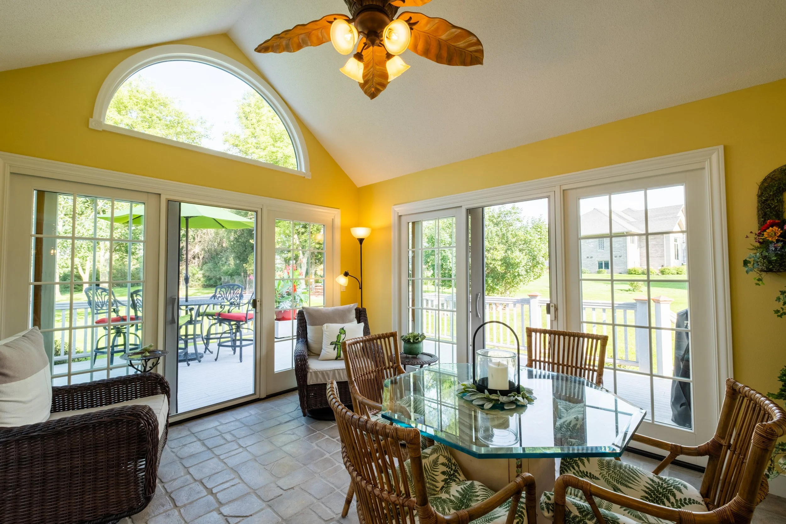 Bright sunlit room with large windows and sliding glass door leading to a porch. There is a glass dining table with wicker chairs, a wicker sofa with cushions, and a ceiling fan with leaf-shaped blades. Outside, patio furniture and lush green trees a