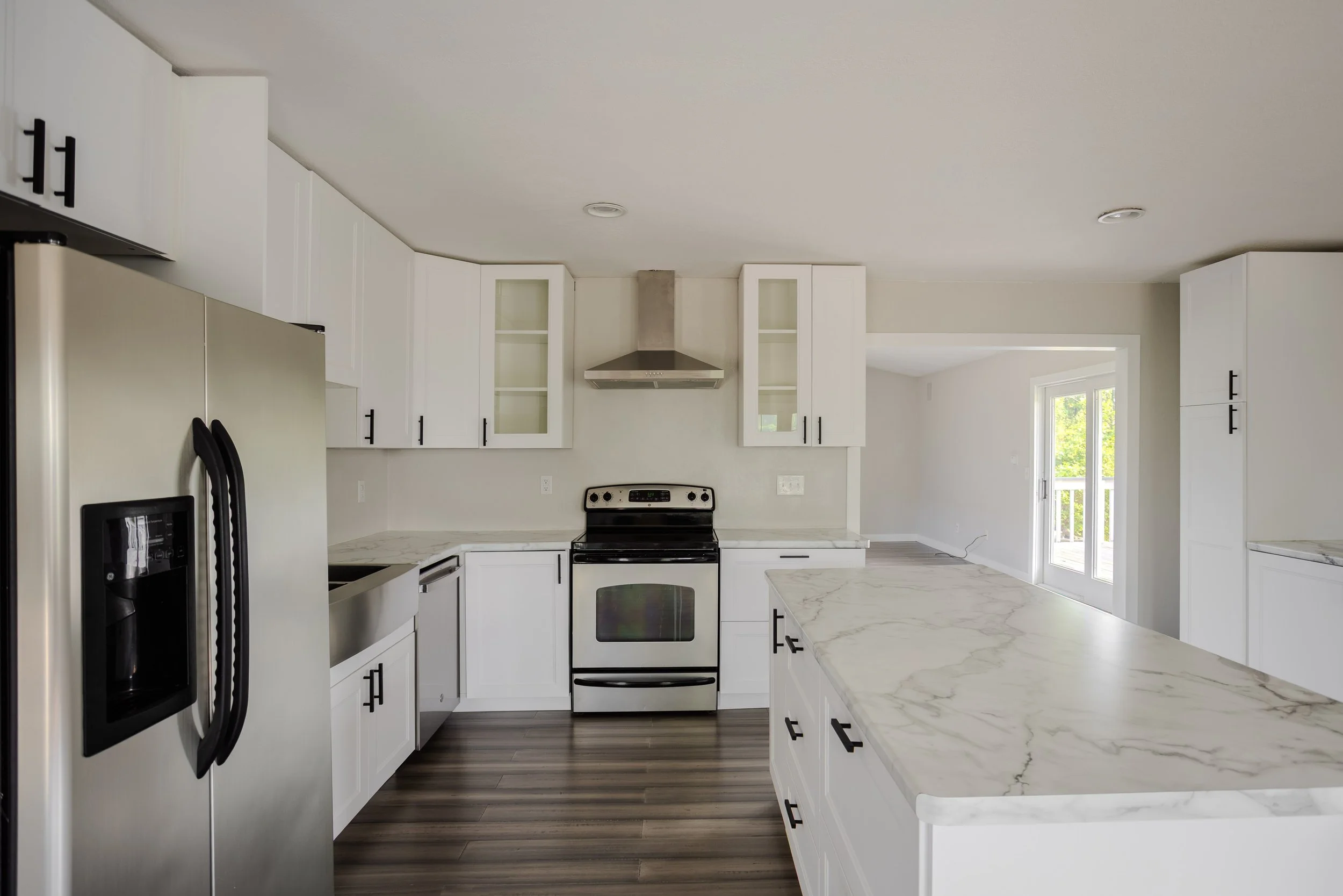 Modern white kitchen with marble countertops, black cabinet handles, stainless steel refrigerator, oven, and hood vent, with hardwood flooring and a door leading outside