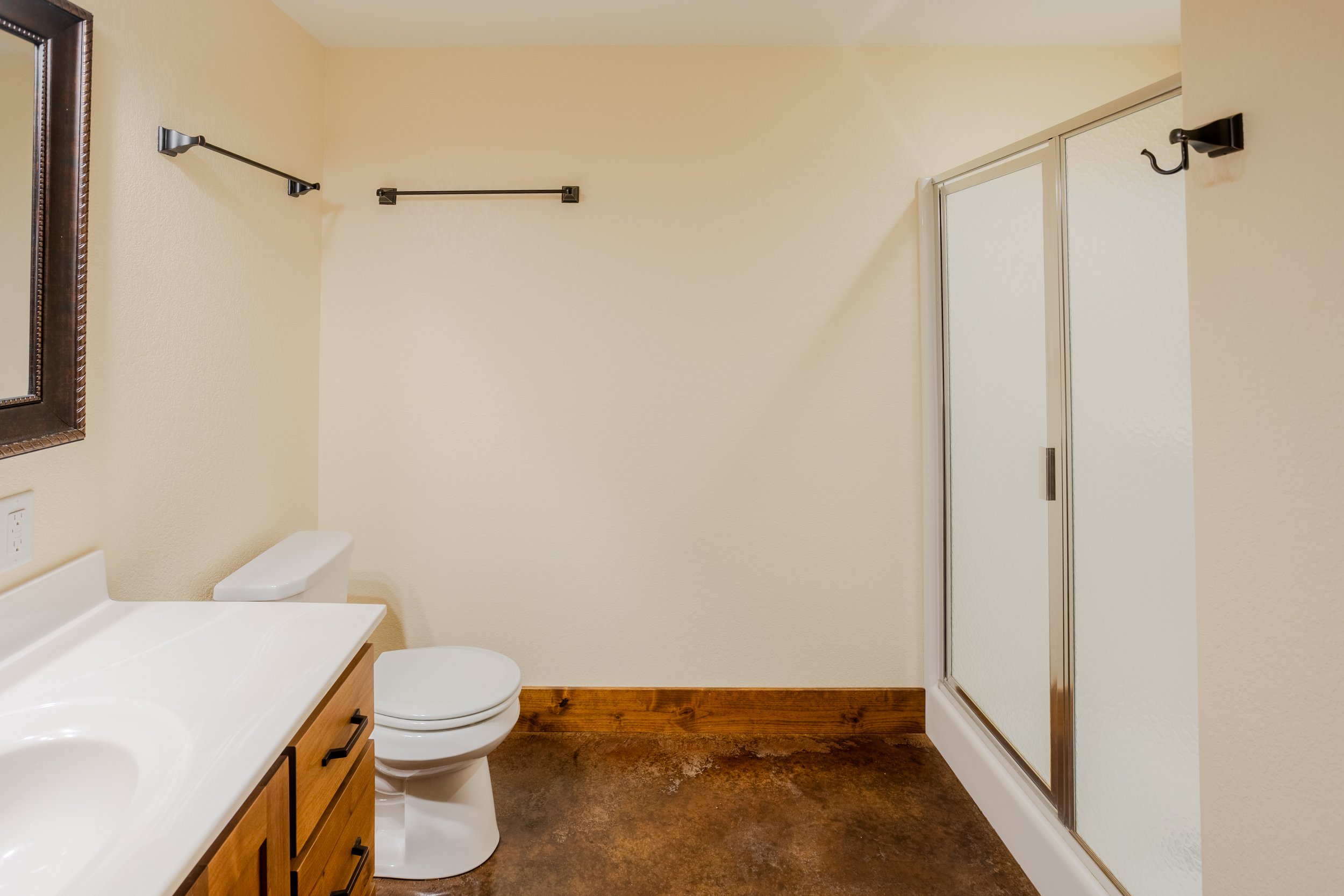 Bathroom with a white countertop and sink, wooden cabinet drawers, a toilet, a large mirror with a wooden frame, yellow painted walls, a sliding glass shower door, two black hooks on the wall, and hardwood flooring.