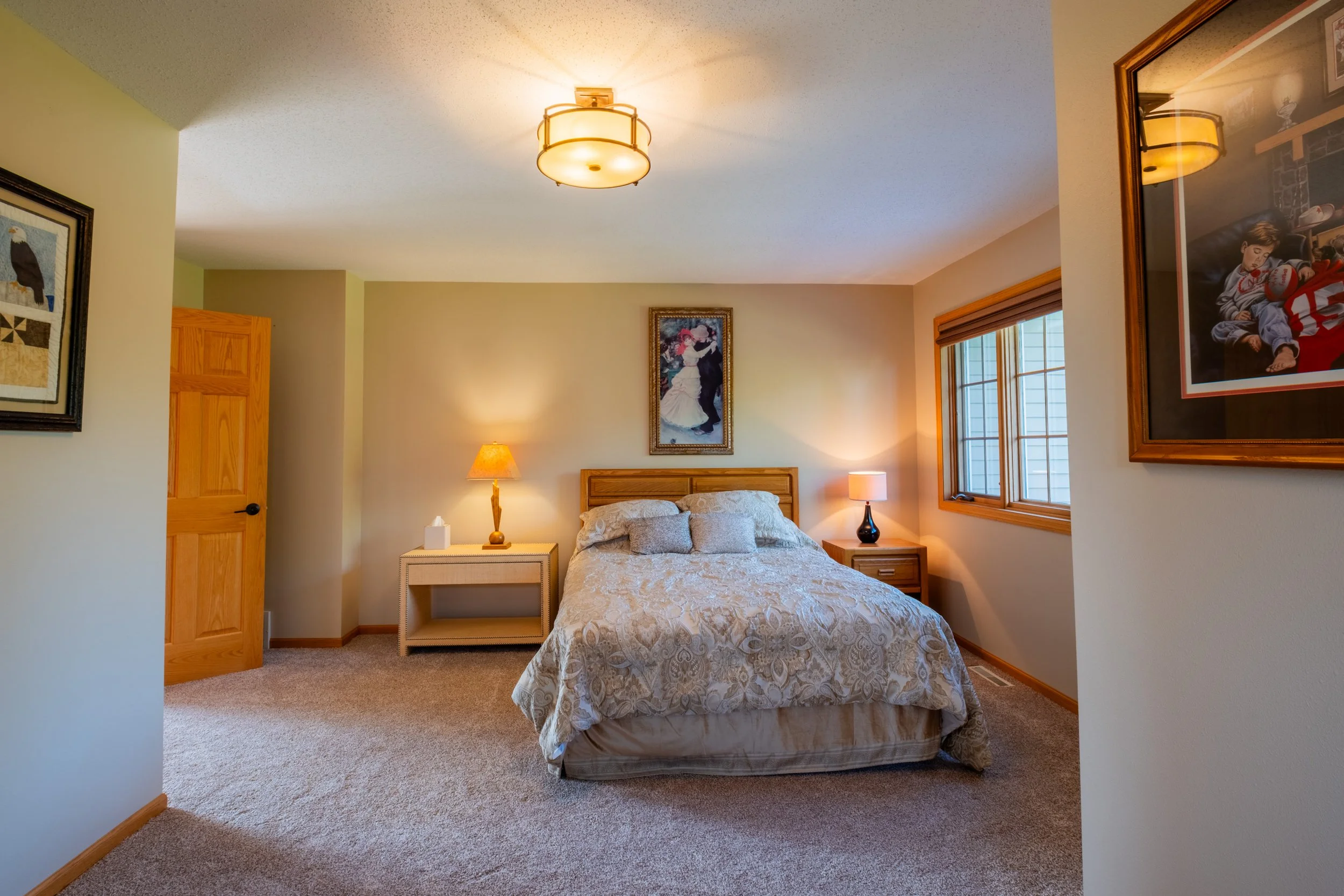 A cozy bedroom with a bed covered in a patterned bedspread, two bedside tables with lamps, framed artwork on the walls, a window with wooden blinds, and a ceiling light fixture.