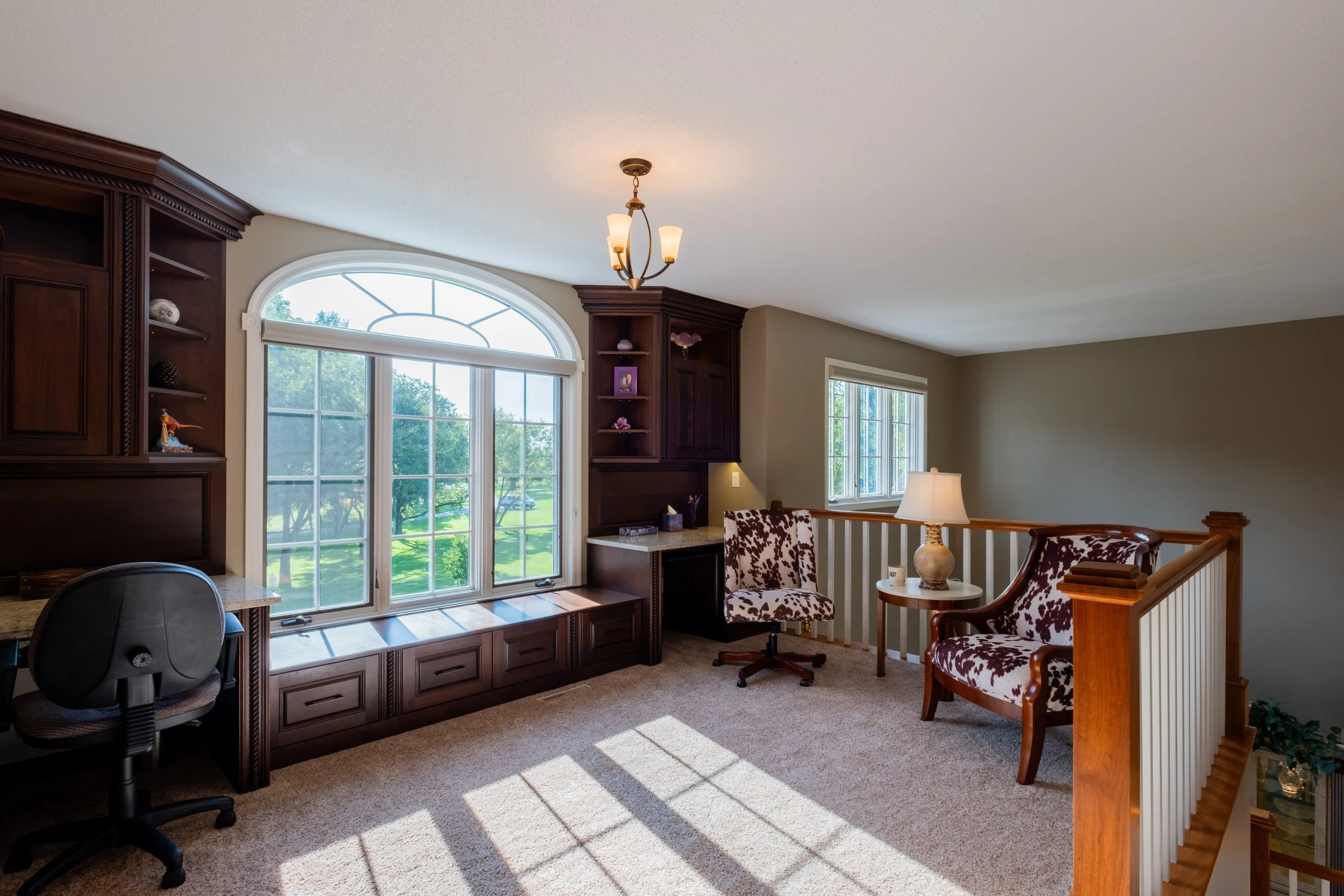 Living room with large arched window, wooden built-in shelves, a chandelier, a desk with a black office chair, and a seating area with two patterned chairs and a side table.
