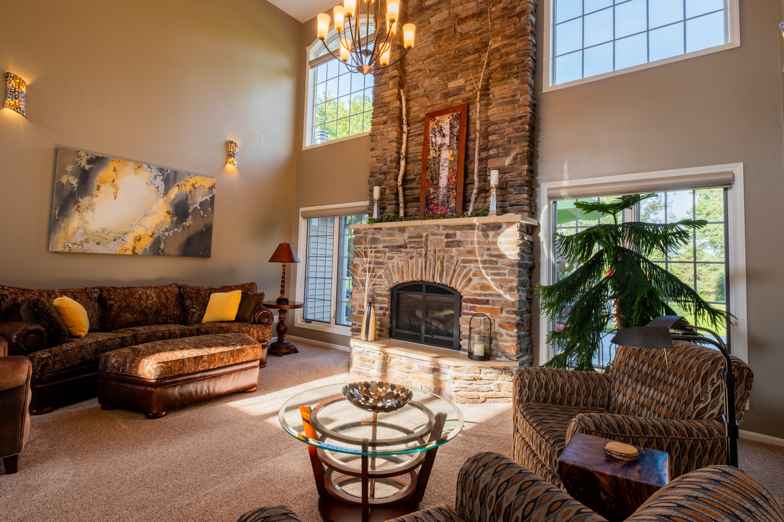 Living room with a stone fireplace, large windows, and various seating including a floral-patterned sofa and armchair, decorated with lamps, artwork, and plants.