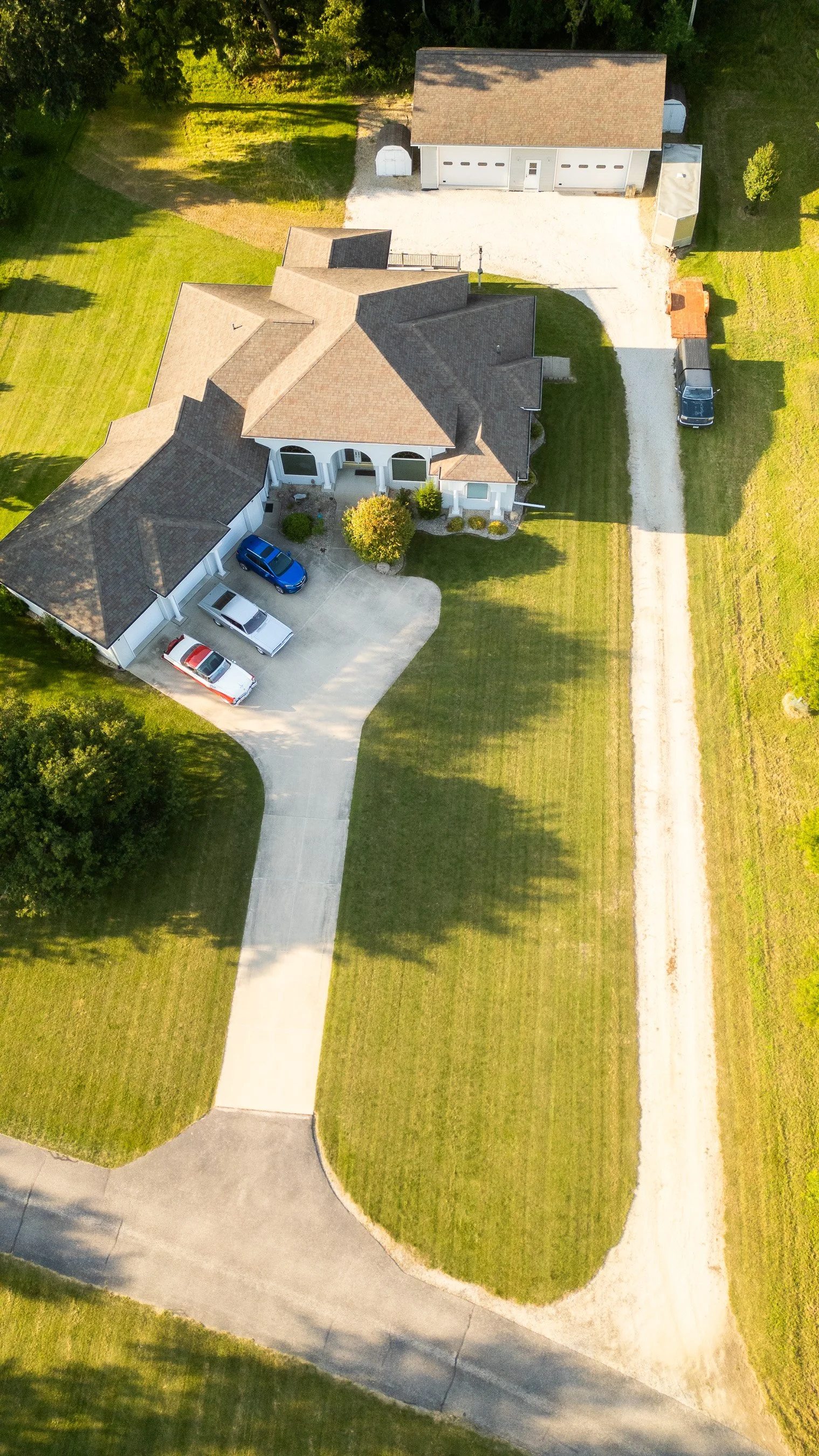 An aerial view of a house with a brown shingle roof, a driveway with three parked cars, and a large grassy yard with trees. A gravel path runs alongside the yard, leading to a detached garage and shed.