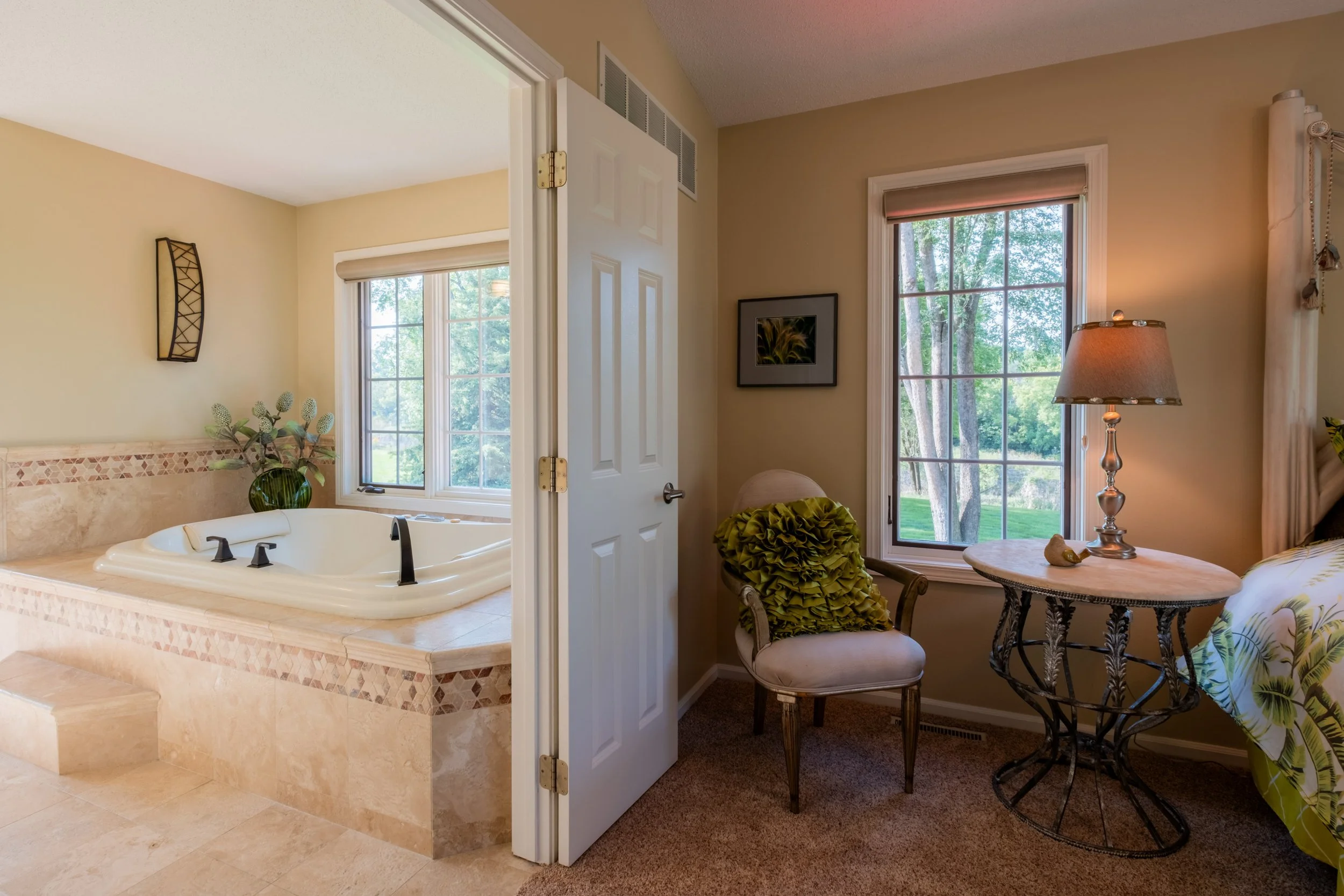 A bedroom with a small sitting area near a window and a bathroom with a large bathtub visible through an open door.