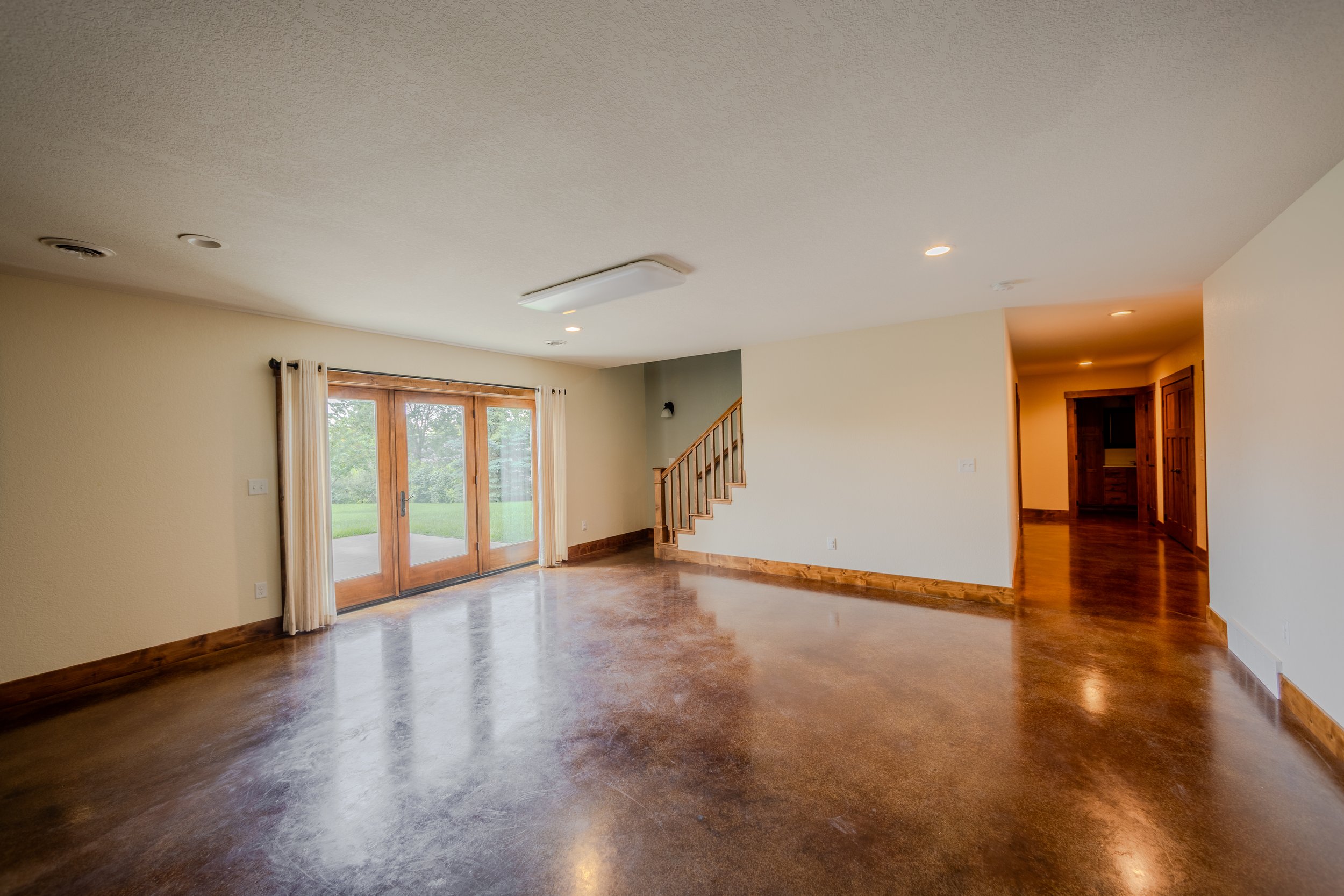 A spacious living room with polished wooden floors, a large sliding glass door with white curtains, and a staircase with wooden railings leading upstairs. There is a door to another room with wooden doors, and the room is well-lit with natural light.