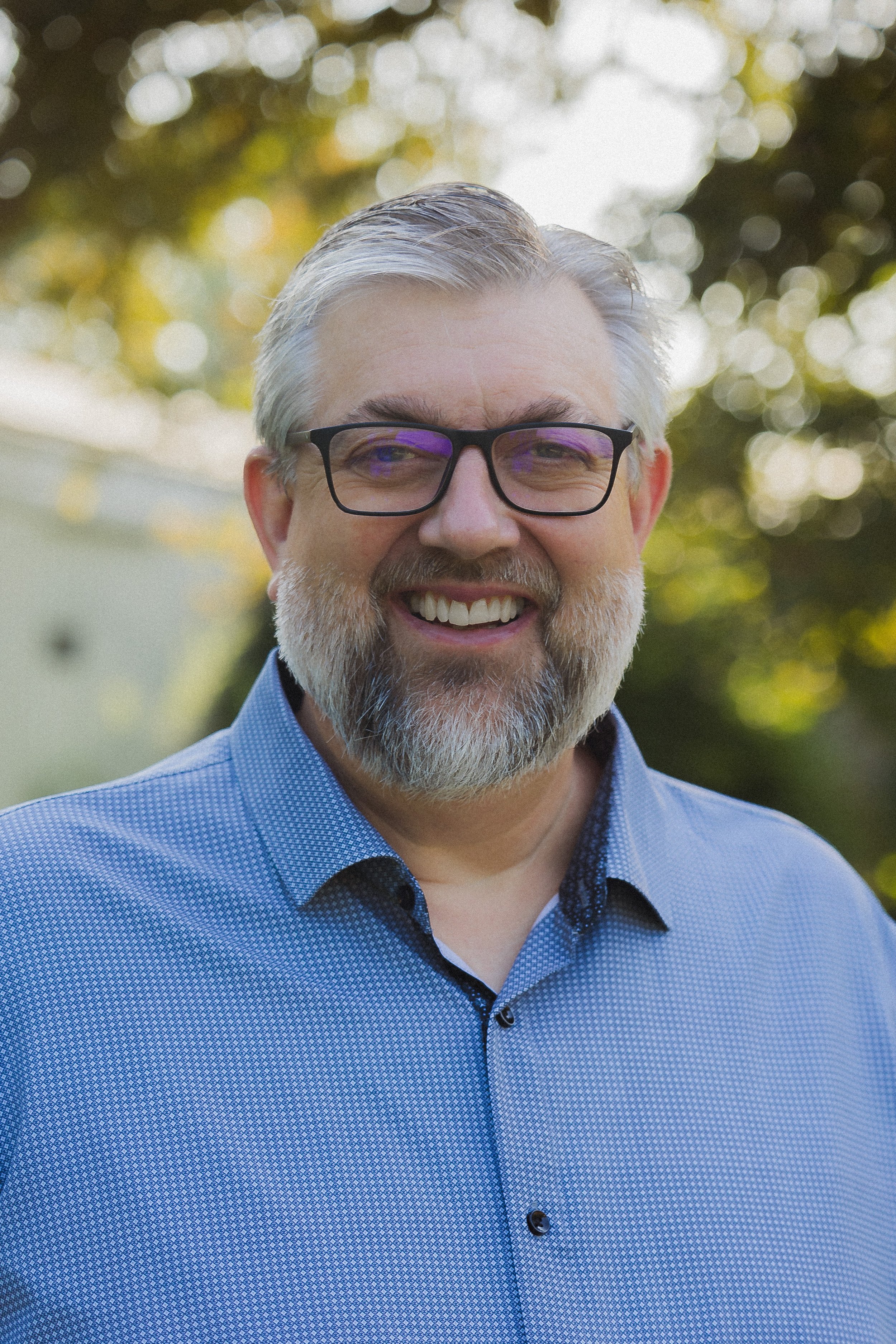 Close-up photo of smiling man with gray hair, beard, wearing glasses and a blue patterned collared shirt, outdoors with blurred trees in the background.
