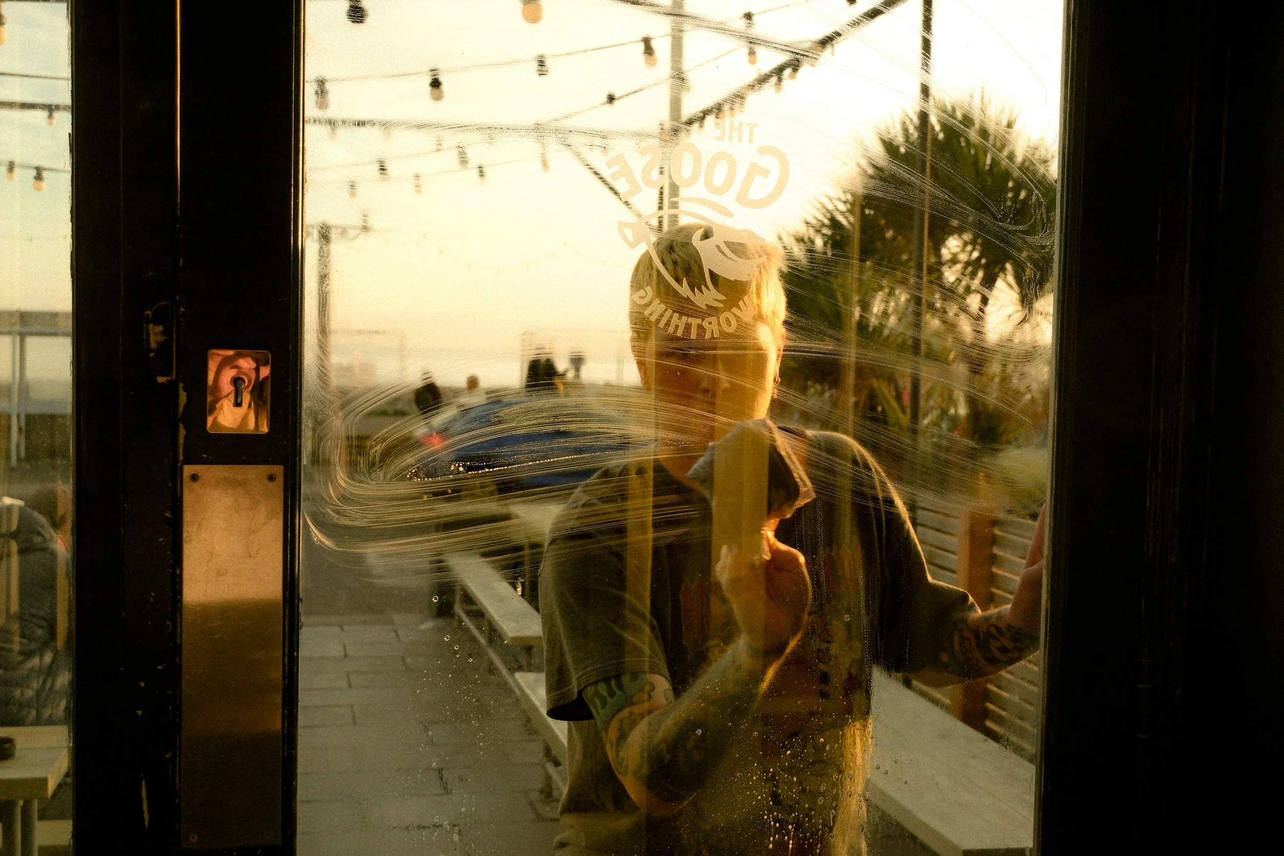 Reflection of a person taking a photo in a glass door, with outdoor string lights, a tree, and a sunset in the background.