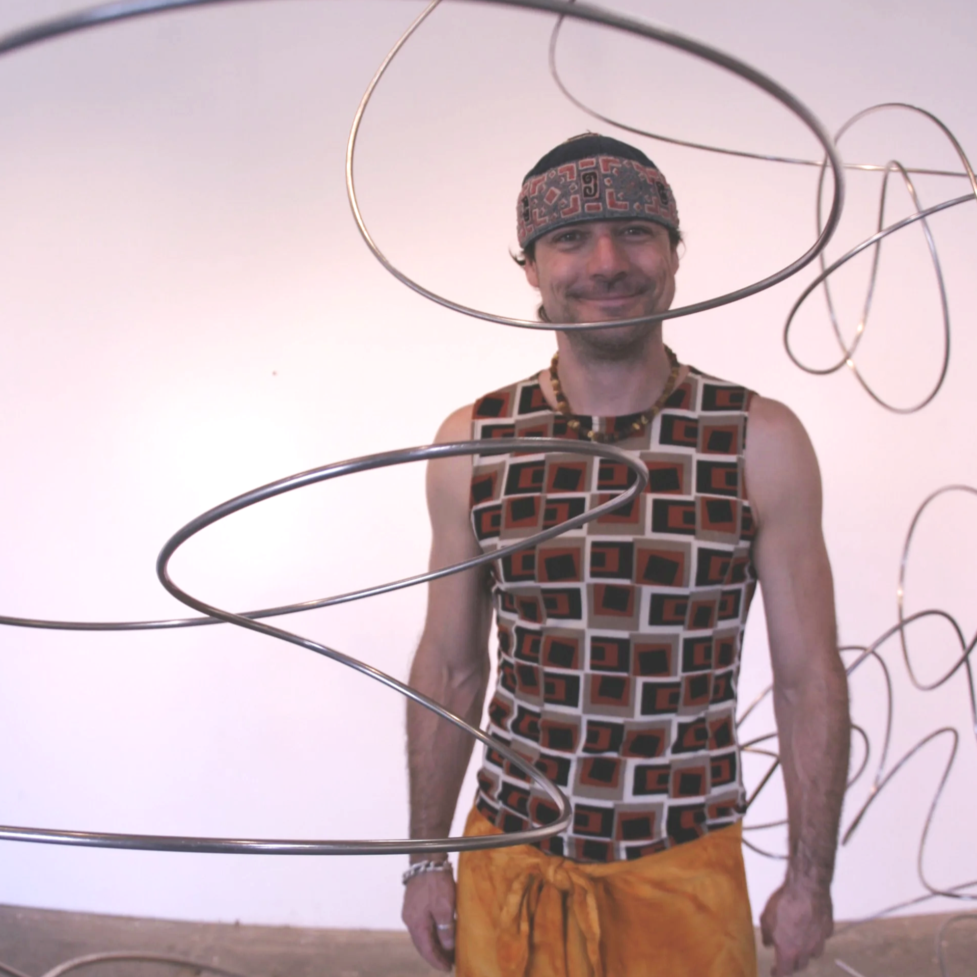 A man in a geometric patterned sleeveless shirt and yellow shorts is smiling and standing against a plain light pink background, with a metallic sculpture in front of him.