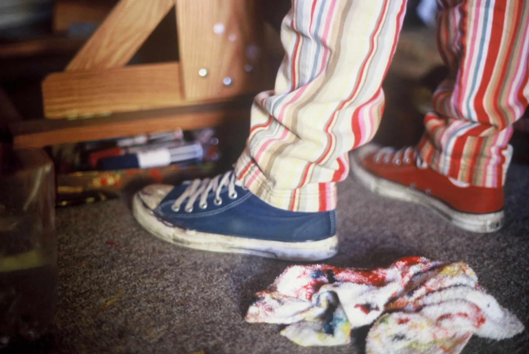 Close-up view of a person's lower body, wearing striped pants and sneakers, standing on a carpeted floor with a colorful cloth or garment on the ground nearby. Part of a wooden piece of furniture is visible in the background.