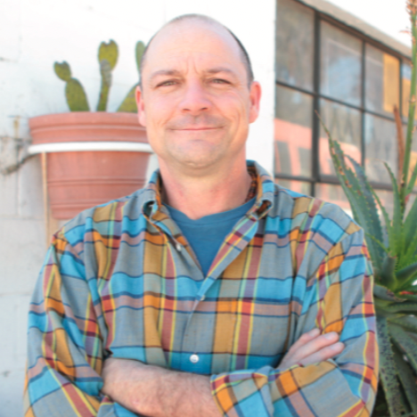 Man standing outdoors with arms crossed, wearing a colorful plaid shirt, with succulents in the background.