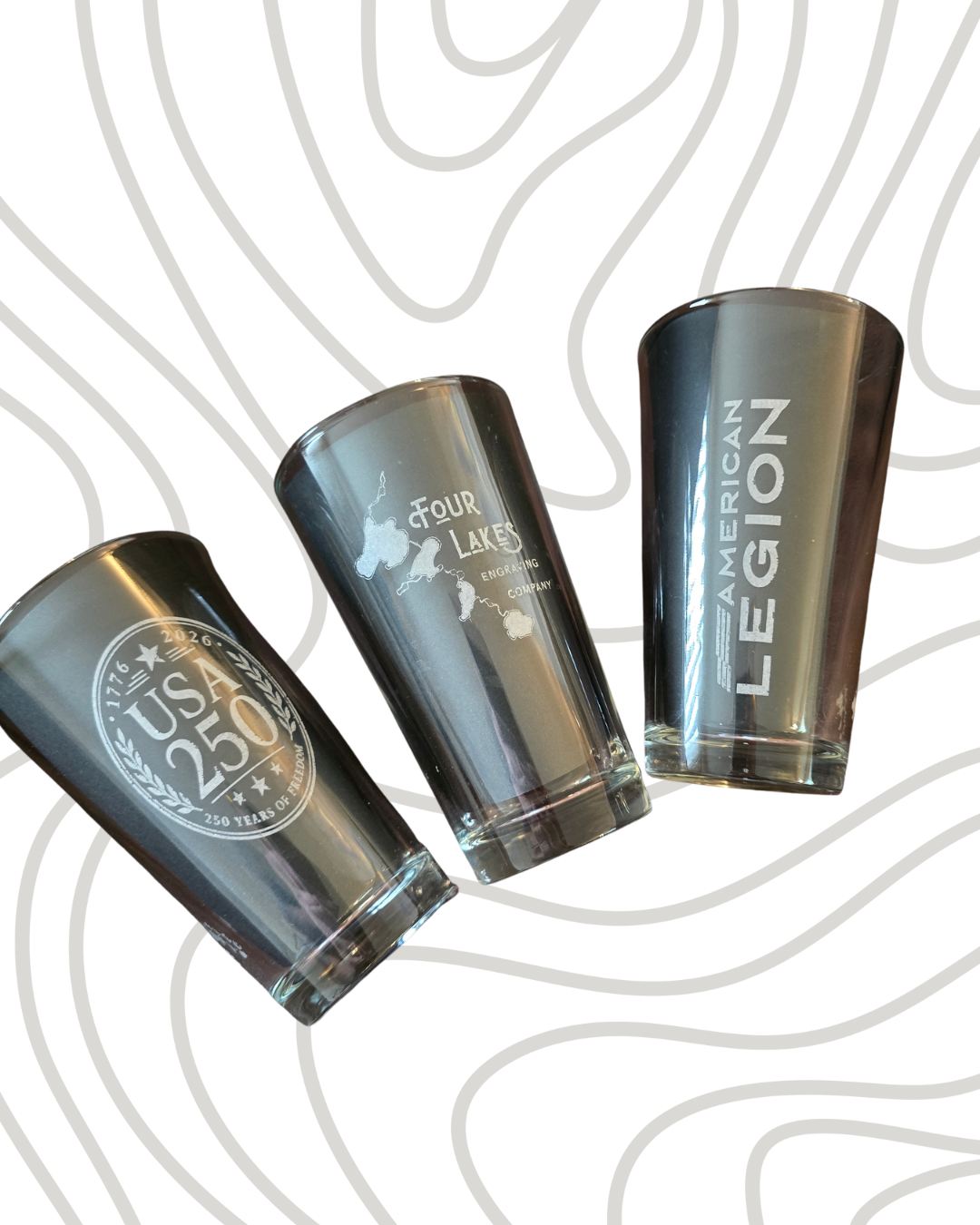 Three engraved whiskey glasses with logos representing USA, Four Lakes, and American Legion, placed on a white background with a topographic map design.