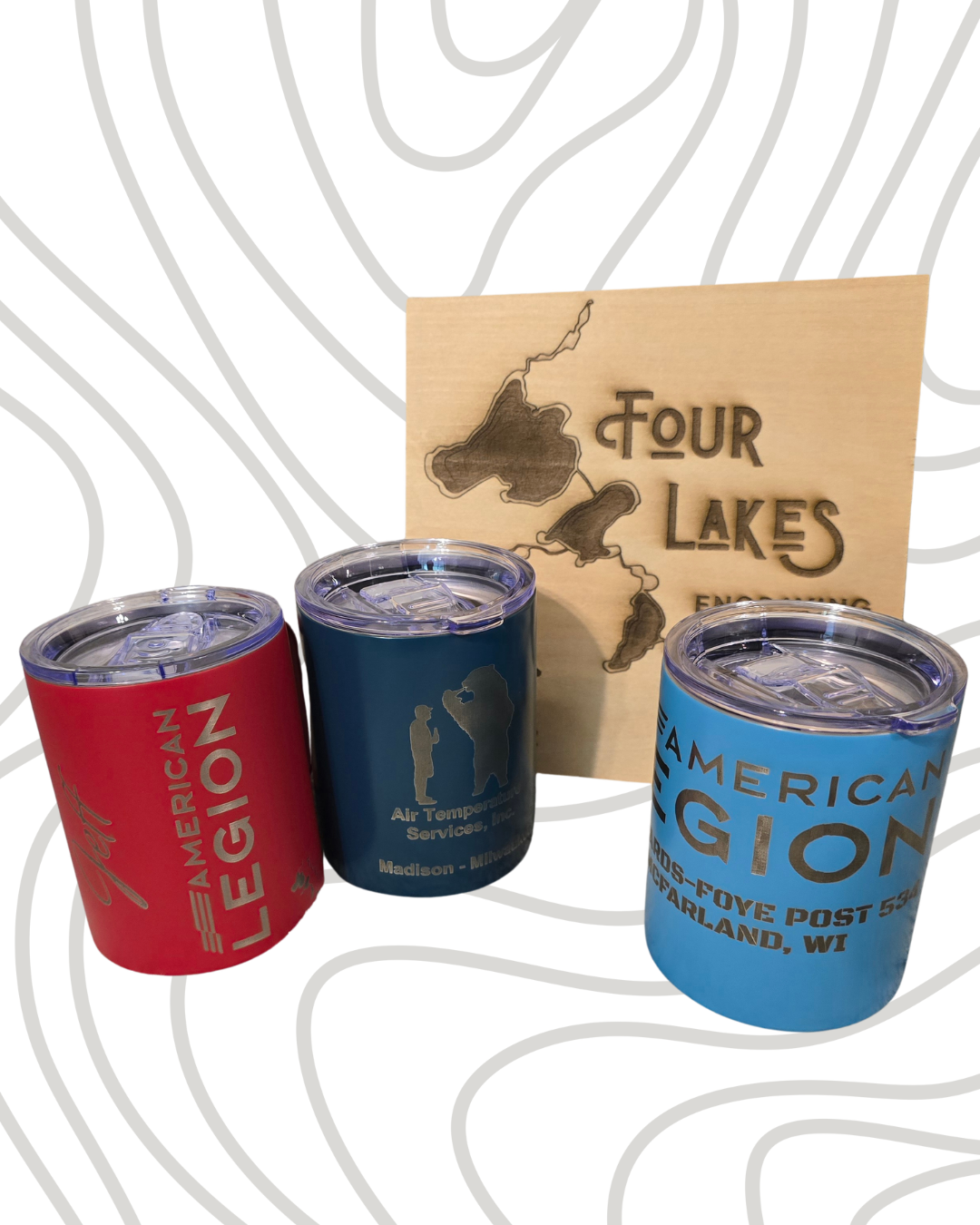 Three insulated travel mugs with metal text and logos, one red, one blue, and one light blue, placed in front of a wooden sign with the map of Four Lakes, Wisconsin. The mugs have designs promoting American Legion and air temperature services.