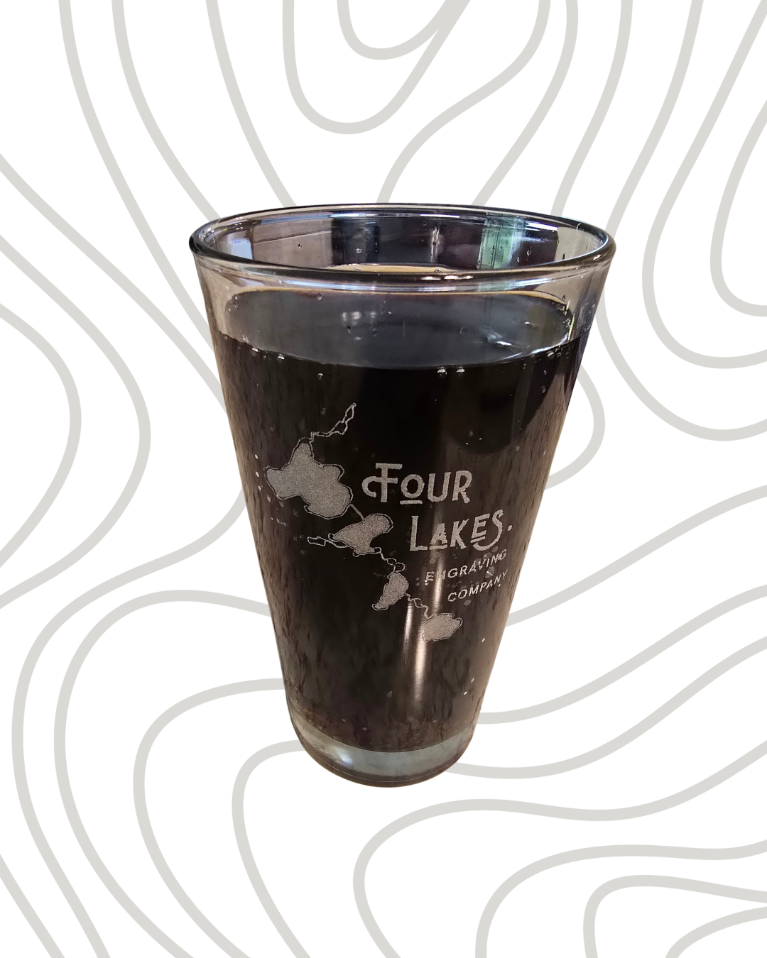 A glass of dark beverage on a white background with a gray topographic pattern, featuring a logo for 'Four Lakes Engraving Company' with a map of four lakes.
